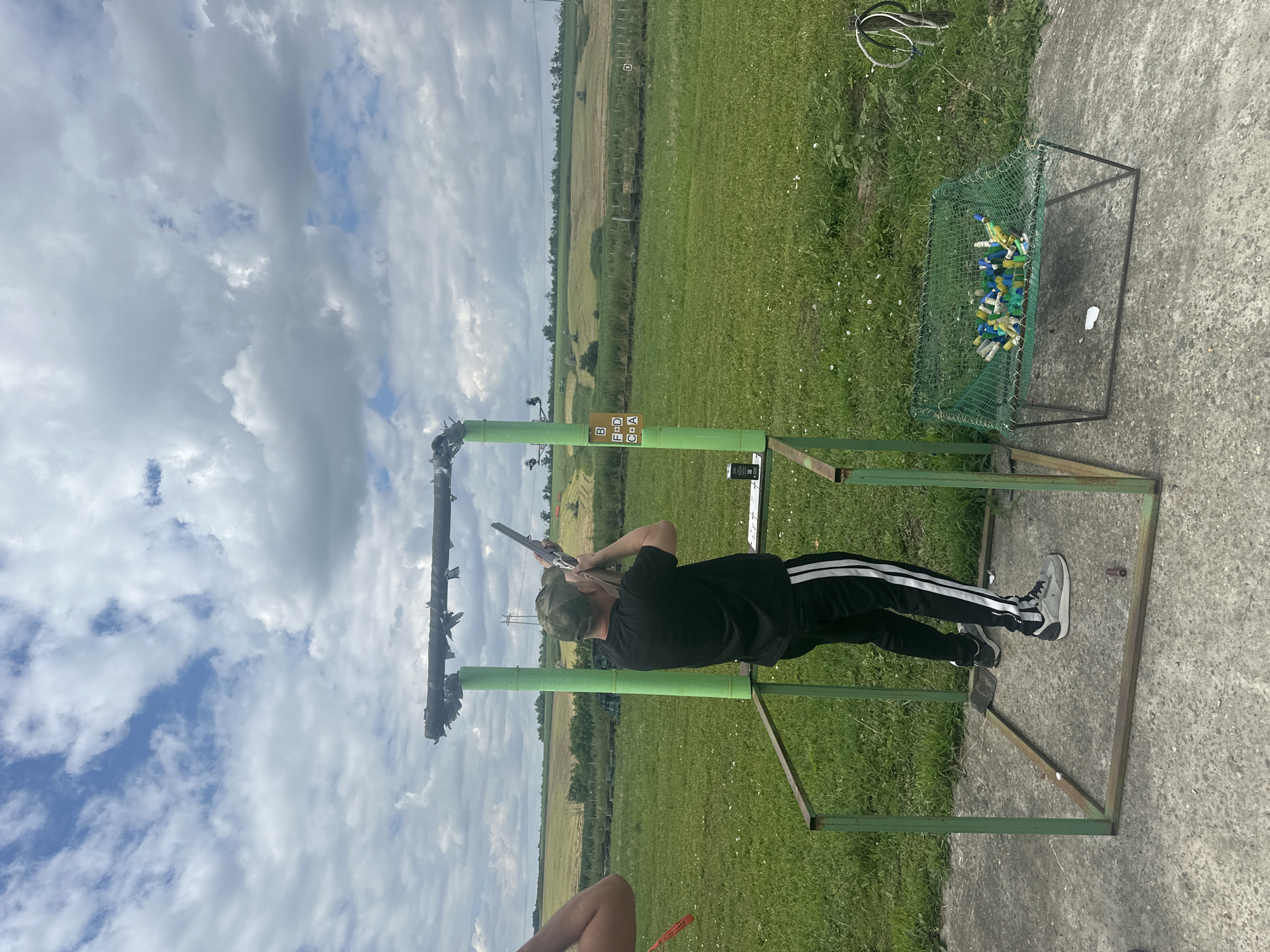 The image captures a person aiming a shotgun at a clay pigeon shooting range. The subject is a young person wearing a black t-shirt, black pants with white stripes, and a green baseball cap. They are positioned within a metal frame structure, holding the shotgun to their shoulder. In the background, a green field stretches towards a distant tree line and agricultural land. The sky is partly cloudy with a mix of blue patches and large, grey clouds.
To the right of the frame, a green metal cage contains numerous colorful clay pigeon targets, mostly blue, yellow, and white. A small, brown sign with letters and symbols is attached to the green metal pole in the foreground. A concrete surface surrounds the shooting stand. The overall scene suggests an outdoor recreational activity under a cloudy daytime sky. The location is identified as Фельдмаршальский, Russia.