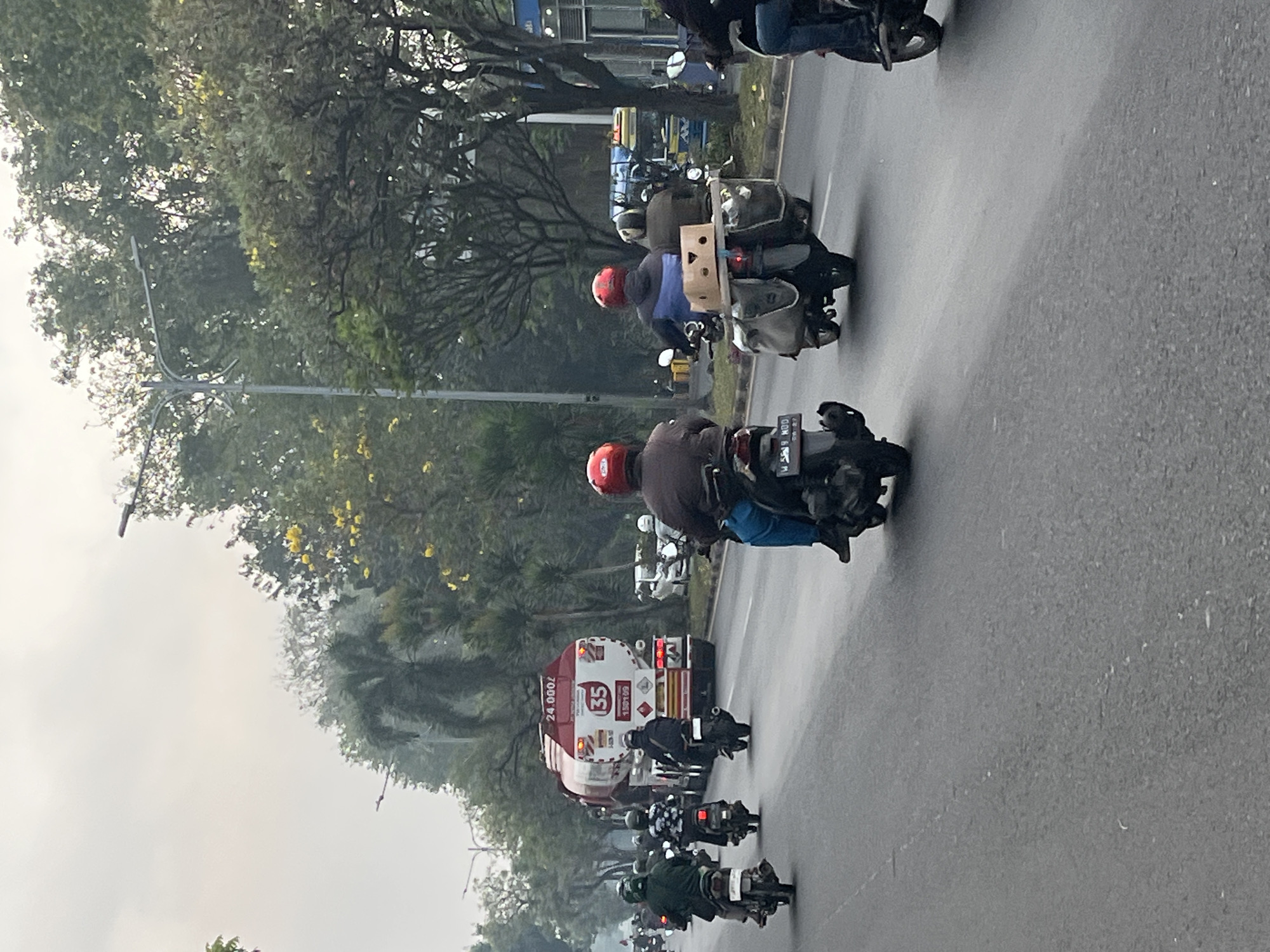The image depicts a busy street scene in Gelam, Indonesia, with a focus on traffic, predominantly motorcycles. In the foreground, several individuals on motorcycles are visible, some with passengers. The road appears to be paved, with lane markings. A large white and red tanker truck with the number "35" and "24.000/" visible on its side is a prominent feature in the middle ground, traveling in the same direction as the motorcycles. Trees line the sides of the road, with some yellow flowers blooming on one of the trees, suggesting a temperate or tropical climate. The sky is overcast, indicating possibly cloudy or hazy weather conditions. The overall atmosphere suggests a typical day in a city with significant motorcycle traffic.