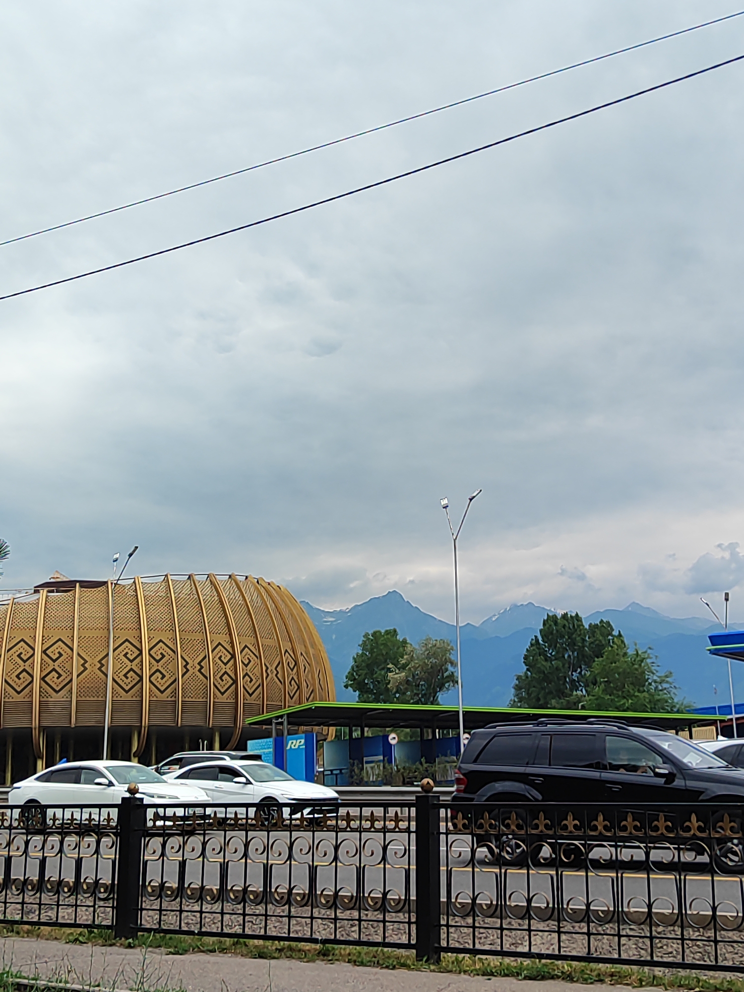 The image depicts a cloudy day in Almaty, Kazakhstan, with a view of the Tian Shan mountains in the background. In the foreground, a black SUV is driving on a road behind a decorative black metal fence. To the left of the SUV, two white cars are parked. A large, golden, domed structure with intricate geometric patterns is visible behind the cars. In the background, several tall green trees stand in front of the mountain range, which has snow-capped peaks. Two streetlights are also present in the scene.