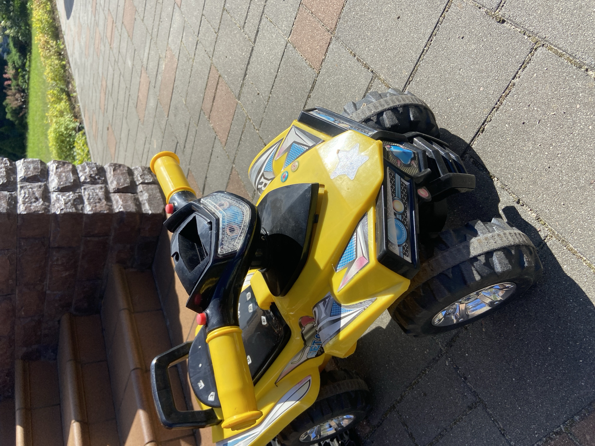 A yellow toy ATV is parked on a paved driveway in front of a brick wall and brown steps. The ATV has black tires with chrome rims and yellow handlebars. The scene appears to be outdoors on a sunny day, with green grass and bushes in the background. The location is Зимородок Днп, Russia.