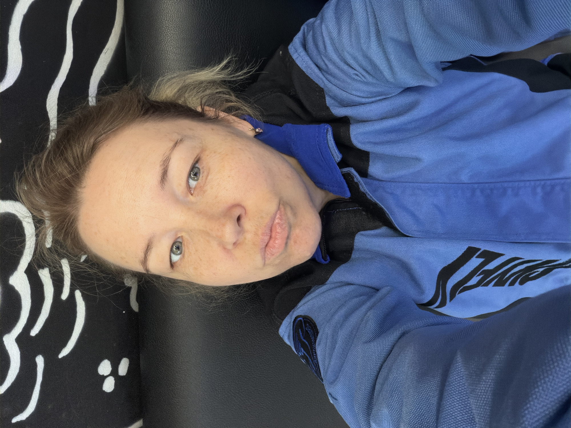 A woman lies on a black and white patterned surface, possibly a couch or bed. She is wearing a blue and black jumpsuit with a logo on the sleeve and lettering on the front. Her hair is blonde, and she has light skin with freckles. The background is out of focus. The location is not discernible from the image.