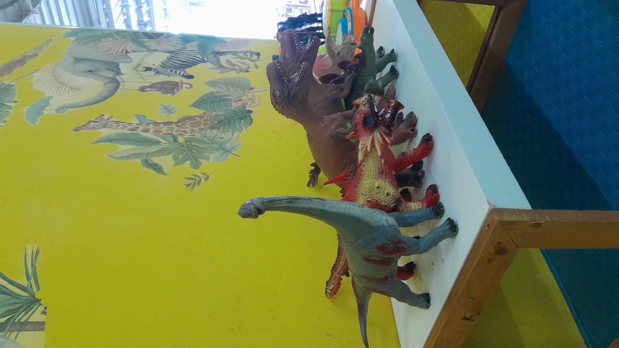 A collection of plastic dinosaur toys are displayed on a white shelf in what appears to be a child's play area or classroom. The toys are arranged in a line from left to right, with a long-necked blue and gray dinosaur in the foreground on the left, followed by a red and yellow spiky dinosaur, a brown Tyrannosaurus Rex, and several other green and brown dinosaurs behind it. The wall behind the shelf is painted bright yellow and decorated with a mural of jungle animals and foliage, including giraffes, elephants, zebras, and monkeys. A wooden frame borders the bottom of the shelf. The floor beneath the shelf is a textured blue mat. The scene is well-lit, suggesting it is daytime. This location is identified as being in the city of Kasiman, Indonesia.
