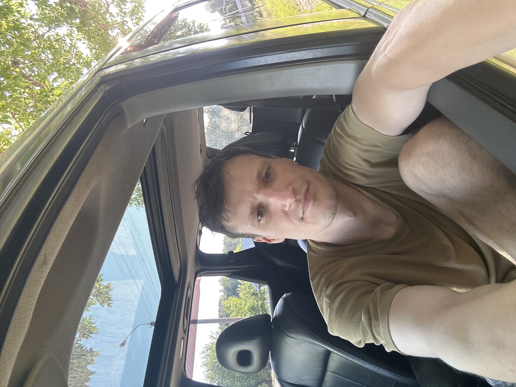 A man is taking a selfie inside a car. He is looking directly at the camera with a neutral expression. The man has short, dark hair and is wearing a plain, light brown t-shirt. He is seated in the driver's seat of a car, with the sunroof open. The interior of the car is visible, including black leather seats. Outside the car, green trees and a blue sky with some clouds can be seen, suggesting a daytime outdoor setting. The background indicates a rural or suburban environment with some vegetation and possibly a fence. The location is identified as Makiv, Ukraine.