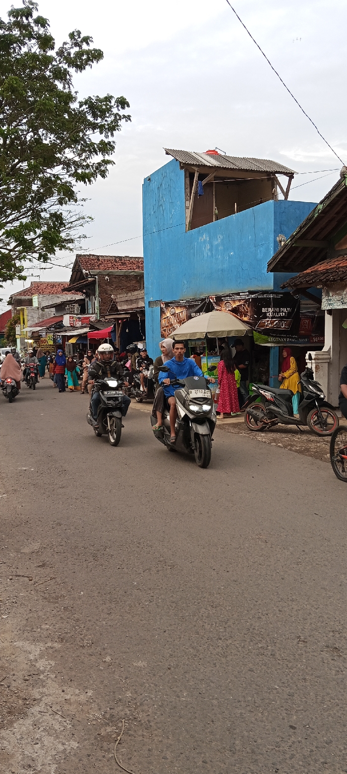 The scene takes place on a paved street in Cangkuang, Indonesia, at the coordinates -7.056 latitude and 107.553 longitude. The street is lined with buildings on both sides, including a blue two-story building with a flat roof. The building features a large sign advertising a product called "MSNUMI." People are walking along the street, including men, women, and children. Some are riding motorcycles. There are several motorcycles parked on the side of the street. The street is relatively empty, and there are no cars present. The weather is clear and sunny. There is a tall tree on the left side of the frame, and a sign with the words "BERANI PILIH KUALITAS" can be seen on the building. A few people stand in front of a store in the background, seemingly engaging in conversation.