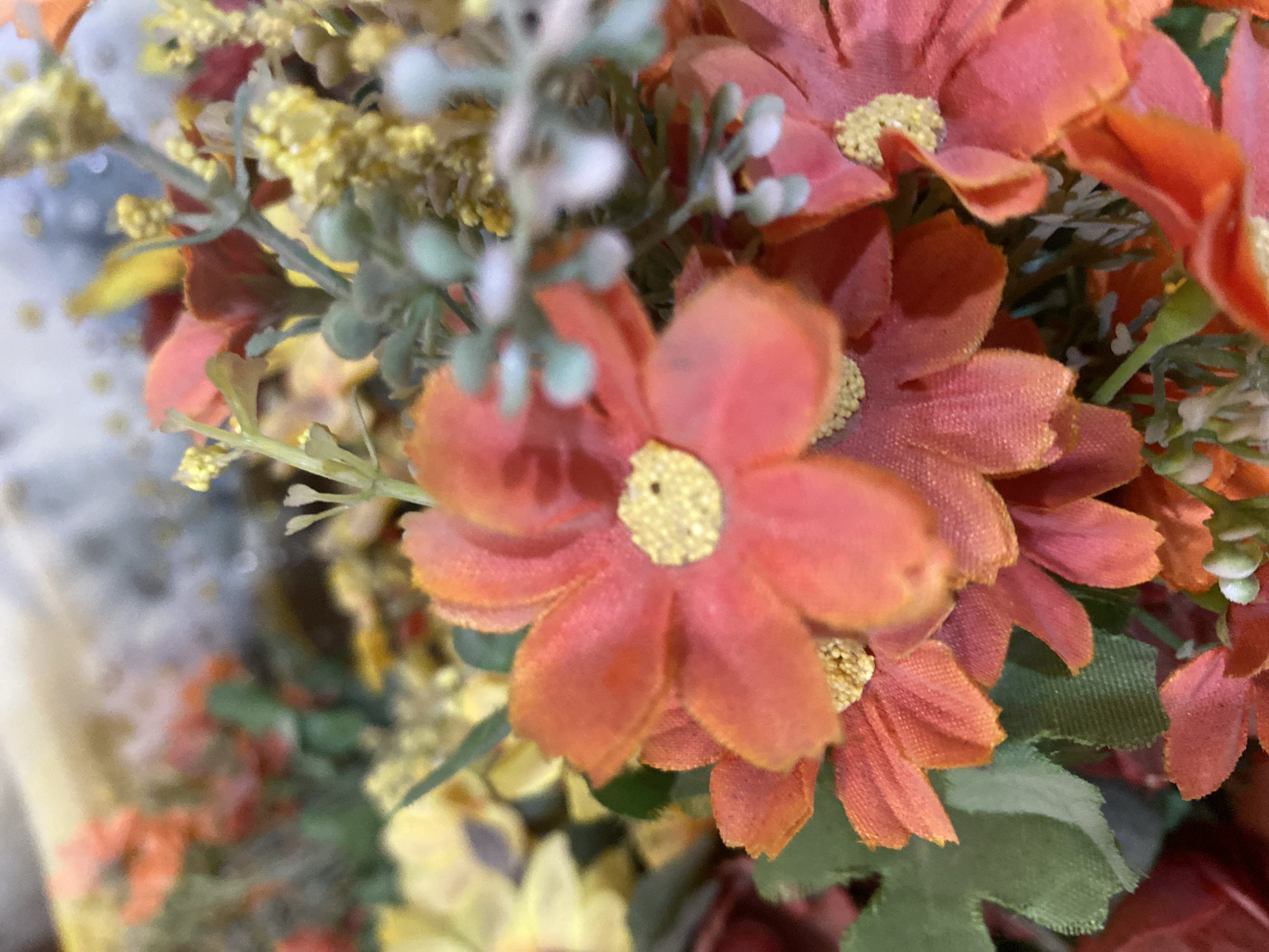 A close-up, slightly blurred, overhead shot displays a bouquet of artificial flowers in warm tones of orange, yellow, and red. The central focus is on several orange daisy-like flowers, each with delicate, textured petals and yellow centers. Green leaves and stems are interspersed throughout the arrangement, along with clusters of tiny yellow flowers and some fuzzy, pale green buds. The background is out of focus, suggesting a shallow depth of field, with hints of lighter colors that could be other flowers or a decorative element. The lighting is soft, creating a gentle illumination of the floral subjects. The scene is devoid of people and action, presenting a still-life composition. This image does not depict Giginyu, Nigeria, or any specific urban environment.