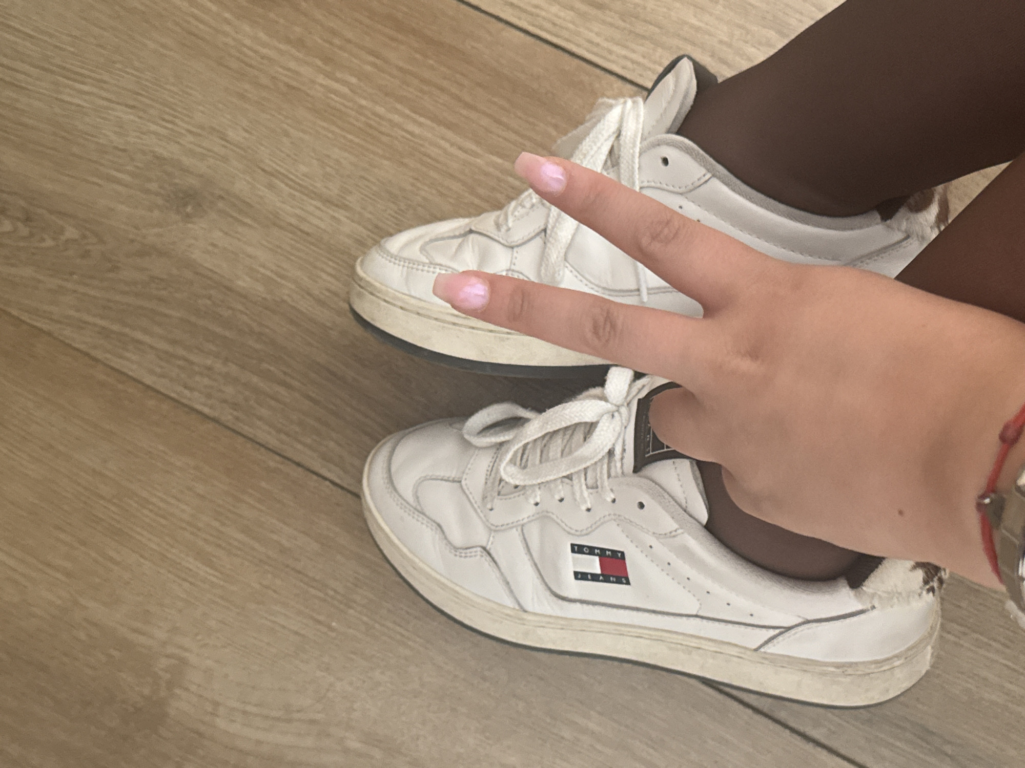 An overhead view captures the lower legs and right hand of an individual positioned on a light brown, wood-grain floor. The person is wearing dark, opaque tights and white low-top sneakers, which show signs of wear, including scuff marks on the soles. The left sneaker displays a rectangular "TOMMY JEANS" logo vertically on its side, featuring the iconic red, white, and blue flag design. The right hand, with light pink iridescent nail polish on its square-shaped nails, is held up in a "V" (peace) sign over the right sneaker. A thin red string bracelet with a small metallic charm is visible on the person's right wrist. The wooden floor planks run diagonally across the frame, with visible seams and some minor surface imperfections. There are no specific visual elements within the image to definitively identify the location as Sofia, Bulgaria.
