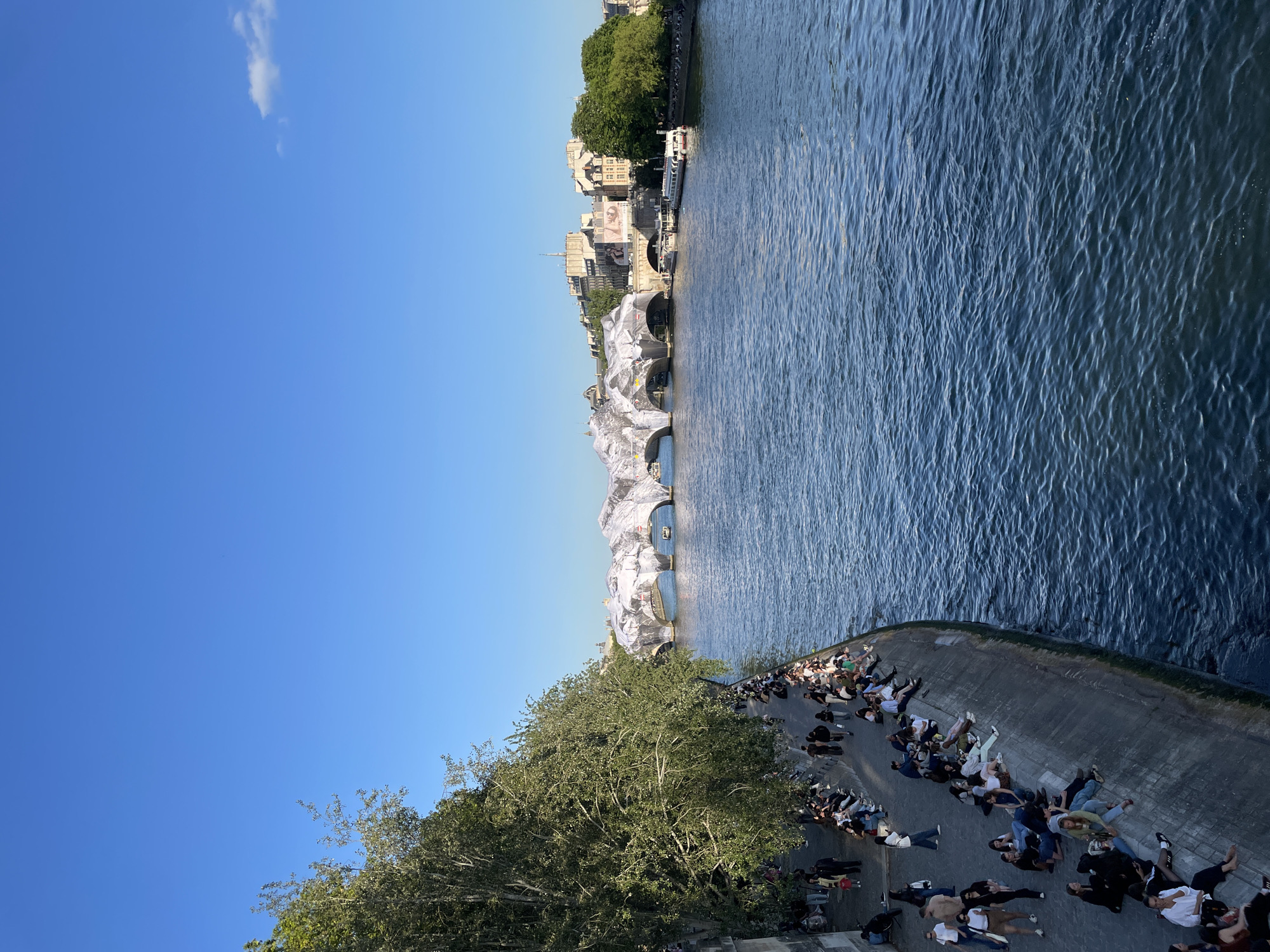 The image shows a portion of the Seine River in Paris, France, featuring the Pont Neuf bridge temporarily wrapped in silvery-white fabric. People are lying and sitting along the concrete embankment near the water's edge, appearing to relax and enjoy the view. There is a tree with full foliage on the left side of the embankment. The river has visible ripples and reflections. Buildings and trees line the opposite side of the river. The sky is clear blue with a single white cloud in the upper left corner.