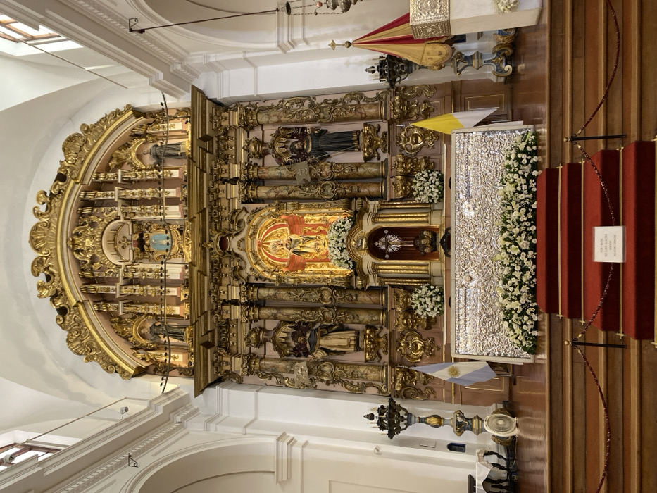 This image depicts the interior of the main altar area of the Basilica and Convent of San Francisco, located in the San Telmo neighborhood of Buenos Aires, Argentina.
The scene is dominated by a grand, multi-tiered Baroque altarpiece, heavily gilded in gold leaf and featuring intricate carvings, columns, and architectural elements. A central, brightly illuminated niche near the top houses a golden statue of a religious figure, possibly the Virgin Mary, with outstretched or winged arms. Below this, at the center of the lower tier, is a smaller, ornate gilded structure, likely a tabernacle, surrounded by arrangements of white flowers. Numerous darker statues of robed figures, possibly Franciscan friars or saints, are positioned within various niches and recesses of the altarpiece.
In front of the altarpiece stands the main altar table, covered by an elaborate, silver-toned metal panel adorned with detailed relief patterns. A dense arrangement of white flowers, including lilies and roses, is placed along the top and front of this altar table.
The area immediately before the altar is a raised platform accessed by three red-carpeted steps. A black rope barrier, supported by metal posts, demarcates this sacred space. A small white sign with black text, reading "POR FAVOR, NO SUBIR AL ALTAR / MANTENER SILENCIO" (Please, do not go up to the altar / Maintain silence), rests on the second step.
To the left of the altar, an Argentine flag stands upright, partially visible, and next to it is a tall, ornate, dark metal candelabrum or lamp stand. A white oscillating fan is visible on the wooden floor near the candelabrum's base. To the right of the altar, a Vatican City flag (yellow and white) is visible, alongside a dark metal stand holding three pointed banners in red and gold fabric.
The surrounding environment consists of white, vaulted ceilings with visible archways and structural elements. High rectangular windows on either side allow natural light into the space. No individuals are present in the image.