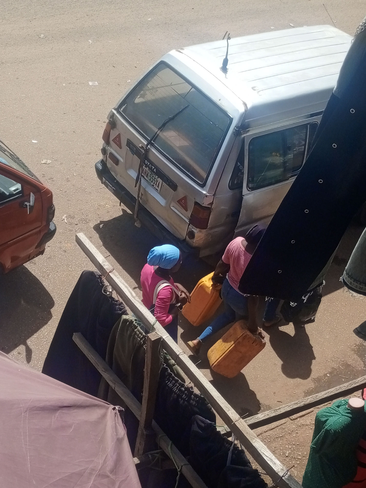 In the city of Bukuru, Nigeria, this image captures a sun-drenched street scene. Two individuals are engaged in transporting water in large yellow jerrycans. One person, wearing a blue headscarf and a pink shirt, is partially visible, holding a jerrycan. The second person, seen from behind and wearing a pink polka-dot shirt and blue jeans, is actively carrying a jerrycan. They are positioned near the rear of a silver Toyota minitruck. The truck has a license plate that reads "ANW 555XA". To the left of the scene, the rear of an orange vehicle is visible. A wooden drying rack with several garments hangs in the foreground, partially obscuring a pink tarp. The ground is paved with a gravelly material, and shadows indicate the strong overhead sun. The overall environment suggests a daily life activity in a Nigerian urban setting.