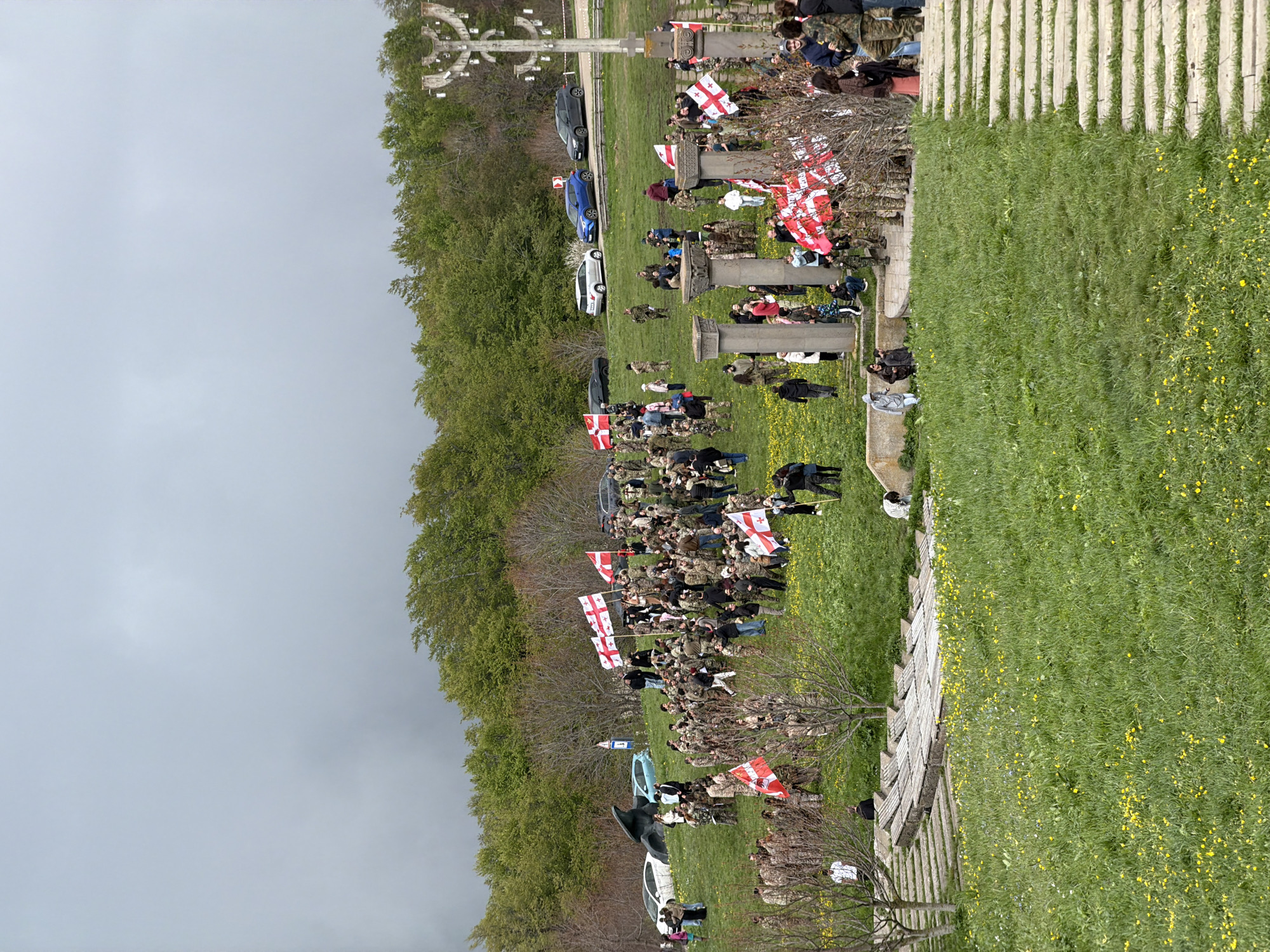The image shows an outdoor gathering in Zemo Nichbisi, Georgia. A large group of people, some in camouflage clothing, are gathered on a grassy area. Many are holding flags with the Georgian cross. Several vehicles are parked nearby, including cars in white, blue, and black. There is a large stone cross monument in the background. To the side of the gathered people, there are rows of concrete columns along a raised grassy area with yellow flowers. Trees are present along the edge of the scene. The sky is overcast.