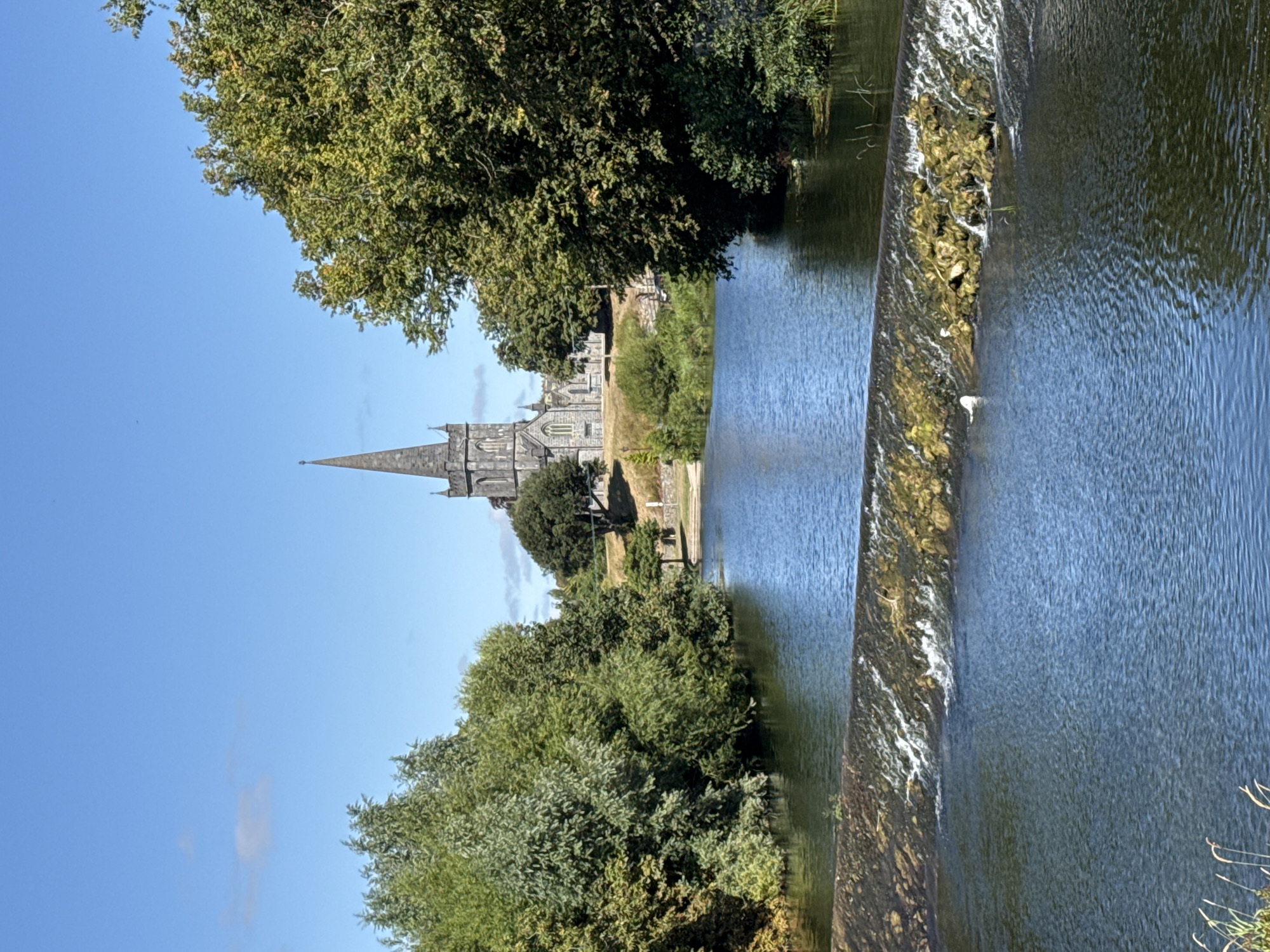 A wide, blue sky with a few wispy clouds is above a lush green landscape in Cahir, Ireland. In the middle ground, a stone church with a tall, pointed spire stands on a hill, surrounded by trees. In the foreground, a river flows, with a low stone weir creating a small waterfall. Trees line the banks of the river, creating a natural frame for the scene. The overall impression is one of peaceful, bucolic scenery.