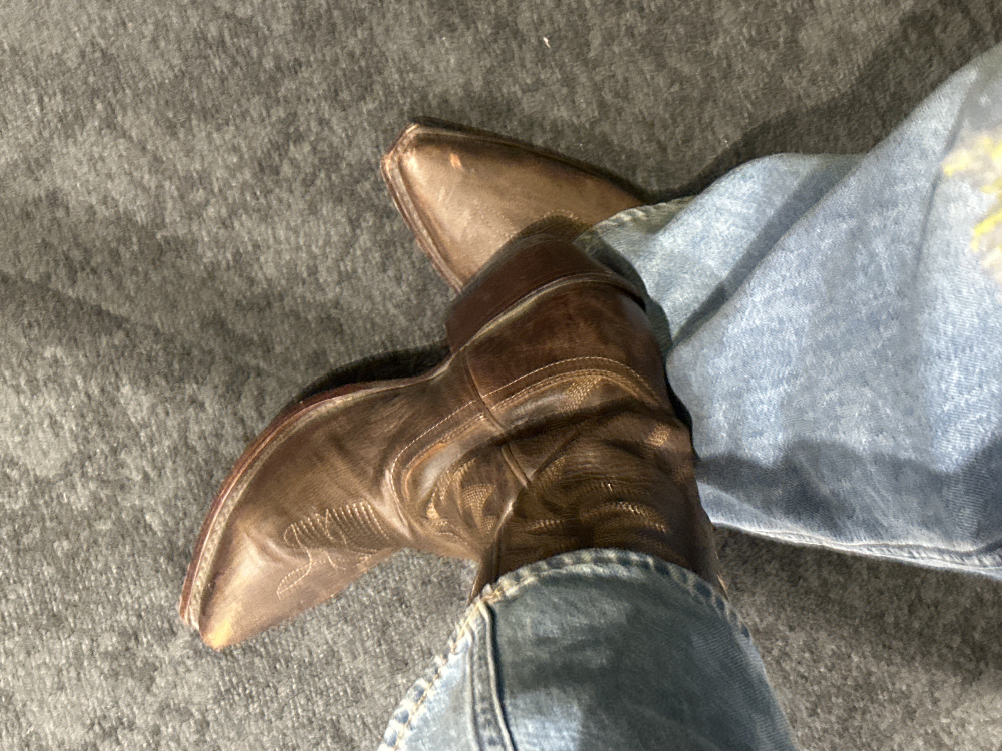 A close-up, high-angle shot shows a pair of brown leather cowboy boots being worn with blue denim jeans. The boots are pointed-toe with detailed stitching. The jeans appear worn. The surface below the boots and jeans is a textured gray fabric. There are no identifiable features to determine a specific location within Los Angeles, United States of America.