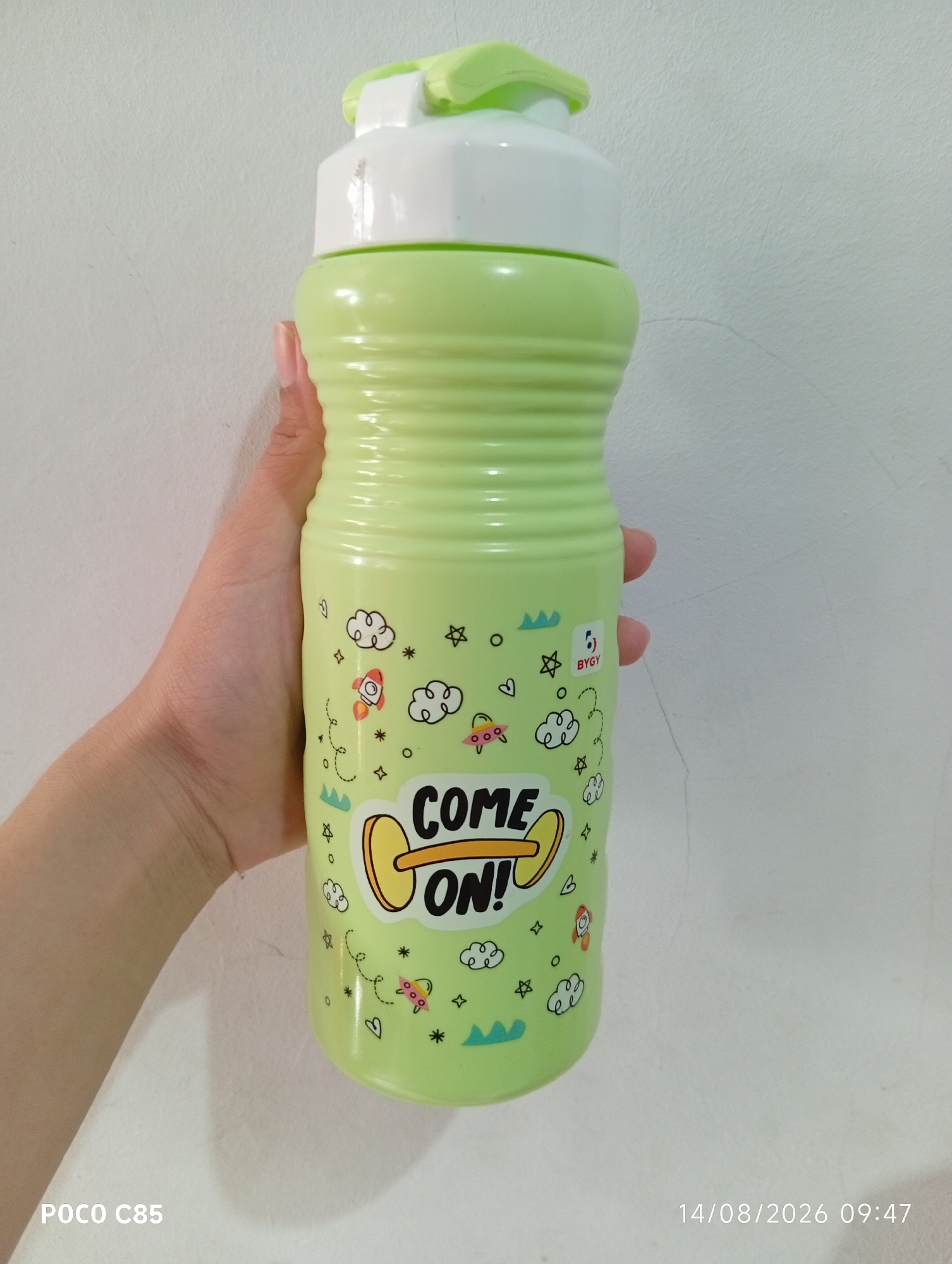 A light green water bottle with a white lid is held by a person's hand against a white, textured wall. The bottle is decorated with cartoon drawings of rockets, clouds, stars, and the text "COME ON!". A watermark in the bottom right corner reads "14/08/2026 09:47". Another watermark in the bottom left corner reads "POCO C85". The environment appears to be indoors.