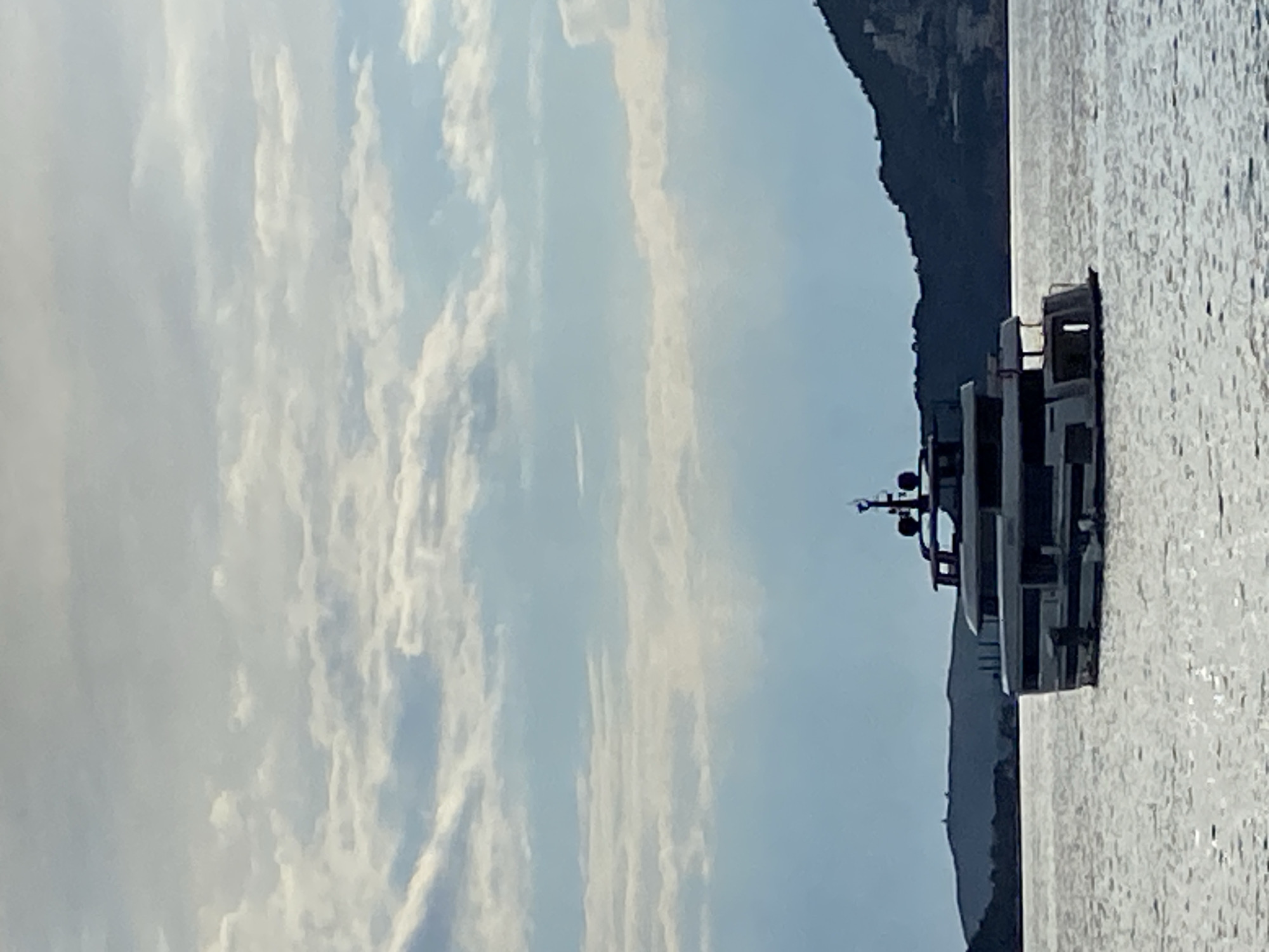 A horizontal image depicts a yacht on water, likely the Adriatic Sea, with a mountainous coastline on the right side and a partly cloudy sky to the left. The yacht is gray and boxy in shape. The water is reflective. The location is probably Pržno, Montenegro based on the geography and coastal features, specifically the mountain shoreline in the background and the presence of yachts in the area. No people are visible.