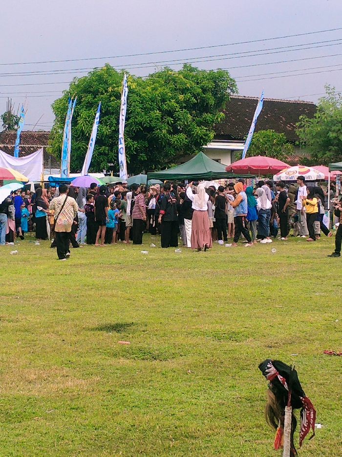 The image depicts a large gathering of people in a grassy field in Karangmalang, Indonesia, at coordinates -7.47 latitude and 111.064 longitude. The group is standing on a field of green grass under a grey sky. There are several blue flags lining the edge of the field. Behind the crowd, there are several stalls with red, white, and blue awnings. In the middle of the field, there is a large group of people, mostly women, with a few men scattered throughout. There is a red and white umbrella on the left side of the image. The people in the crowd are interacting with each other, many of them are smiling and talking. There is a light-colored building behind the stalls. The image was likely taken during a public event or gathering.