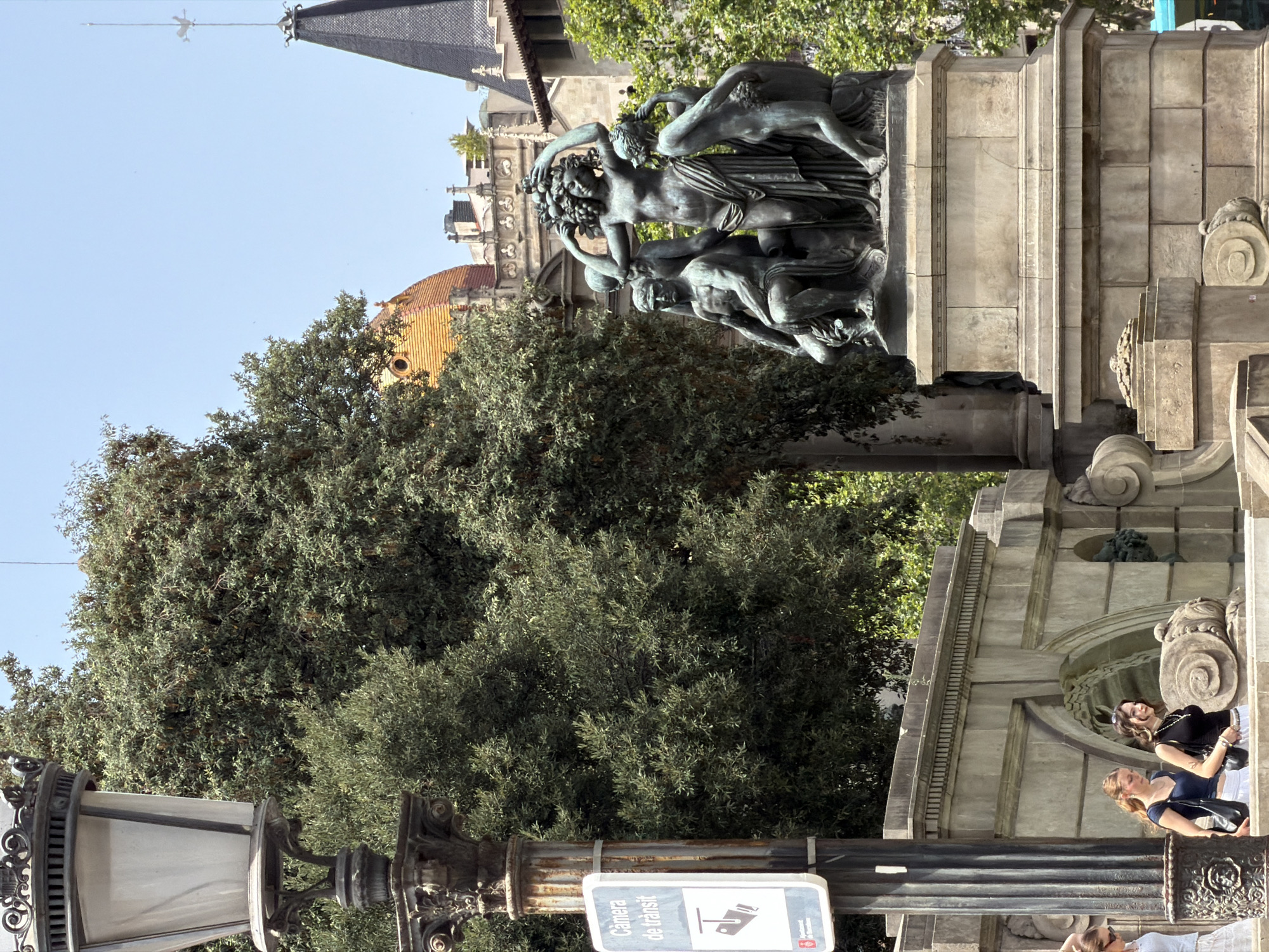 This is a vertical, full-shot photograph of a public monument in Barcelona, Spain, likely taken during the daytime on a clear day. The monument appears to be made of stone and features a bronze sculpture of several human figures intertwined. The sculpture depicts a dynamic scene with figures in various poses, suggesting movement or a narrative.
Behind the monument, a large tree with dense green foliage dominates the middle ground. In the background, architectural elements of buildings can be seen, including a pointed spire on a dark-tiled roof and a section of a building with a curved, orange-tiled roof. The overall setting appears to be a city square or park.
In the foreground, a lamppost with an ornate, dark metal base and a white glass shade is visible. A sign attached to the lamppost reads "Camera de transit," indicating a traffic camera.
Two women are visible near the monument. One woman, wearing a black top and white skirt, is standing and looking to the side. Another woman, in a blue top, is also standing and looking away from the camera. A third person, partially visible at the bottom of the frame, appears to be wearing sunglasses. The people's body language does not convey any strong emotions. The sunlight suggests a bright day.