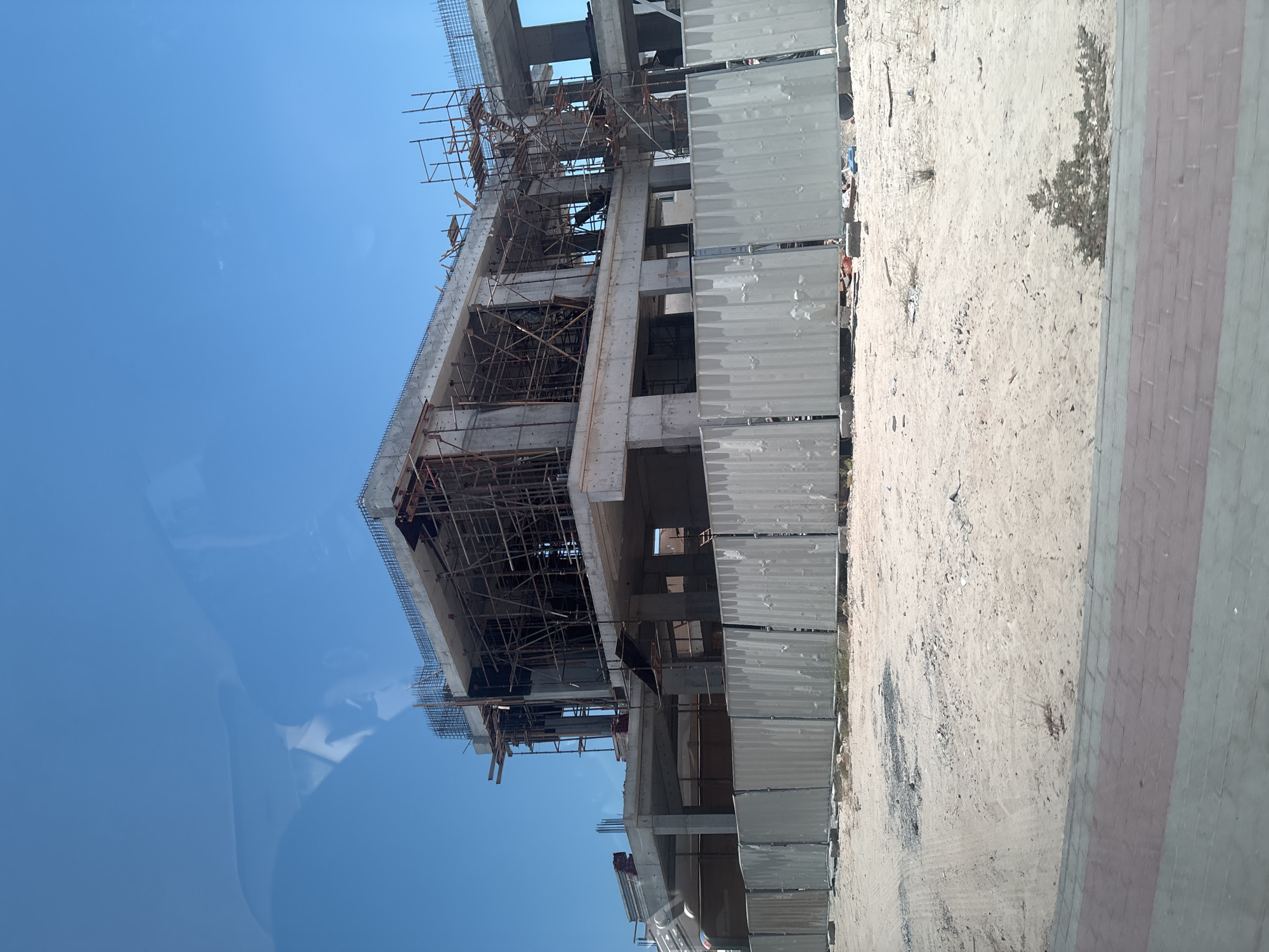 A construction site in Dubai, United Arab Emirates, is depicted under a clear blue sky. The primary subject is a multi-story building under construction, characterized by its concrete frame and extensive scaffolding. The ground surrounding the building is sandy, with some loose debris and sparse vegetation. A corrugated metal fence runs along the perimeter of the construction area, separating it from a paved walkway in the foreground, which features a pattern of brickwork. The image captures the raw, unfinished state of a building project in a typically arid and sunny environment, with no individuals or active construction processes visible.