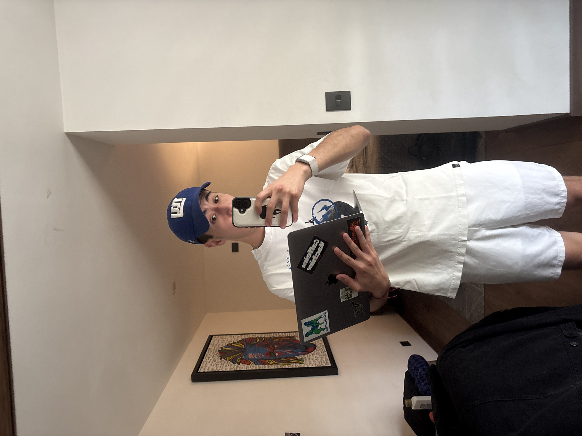 A young man is captured in a selfie taken indoors, appearing to be in a modern home in San Bernardino Tlaxcalancingo, Mexico. The image is rotated 90 degrees counter-clockwise. He stands in what appears to be a well-lit room with light-colored walls and wooden flooring partially visible.
The man, with a surprised or wide-eyed expression, looks directly into the camera. He wears a blue baseball cap with a white logo (possibly an 'F' or stylized 'E'), a white t-shirt with a blue and white graphic design, and white shorts. A light-colored watch or band is on his left wrist, and a thin red string bracelet is on his right. He holds a smartphone with a distinctive white and black patterned case in his right hand, using it to take the photo. In his left hand, he holds an open dark-colored laptop, which has several stickers on its lid, including an Apple logo. The text on the stickers is inverted and partially unreadable; one appears to reflect "delight," and another features a green and white graphic.
In the background, below the man, a framed artwork rests on a white surface, depicting a colorful, mosaic-style head or face, possibly a horse. A dark wall switch or outlet is visible on the wall behind him. To the lower right, a dark jacket or garment is draped over a chair or object, next to a blue, textured item, possibly a cushion or slipper. The overall setting suggests a casual, domestic environment.