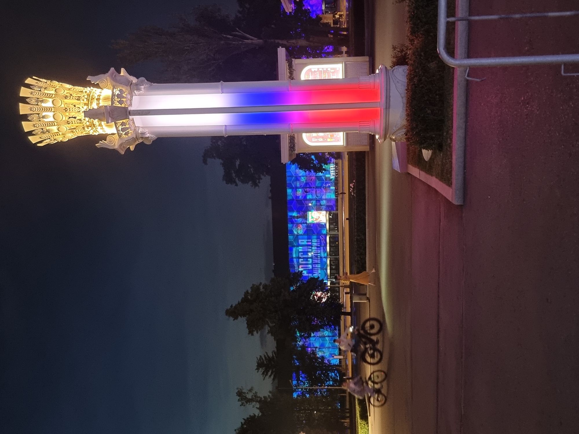 The image shows an outdoor scene at night in Moscow, Russia, likely at an exhibition or park. In the foreground, a tall, ornate lamppost is illuminated with the colors of the Russian flag – white, blue, and red. To the left of the lamppost, two people are riding bicycles on a paved pathway. One person is on a white bicycle and the other is on a dark bicycle. Further in the background, a large building is brightly lit with a blue and purple light show. The building has large windows and the word "РОССИЯ" (Russia) is visible across the facade, along with what appears to be a slogan below it. A woman in a long, flowing yellow dress walks across the pathway in front of the building, between the cyclists and the building. Trees are visible behind the building and to the right of the lamppost. The sky is dark, indicating it is nighttime.