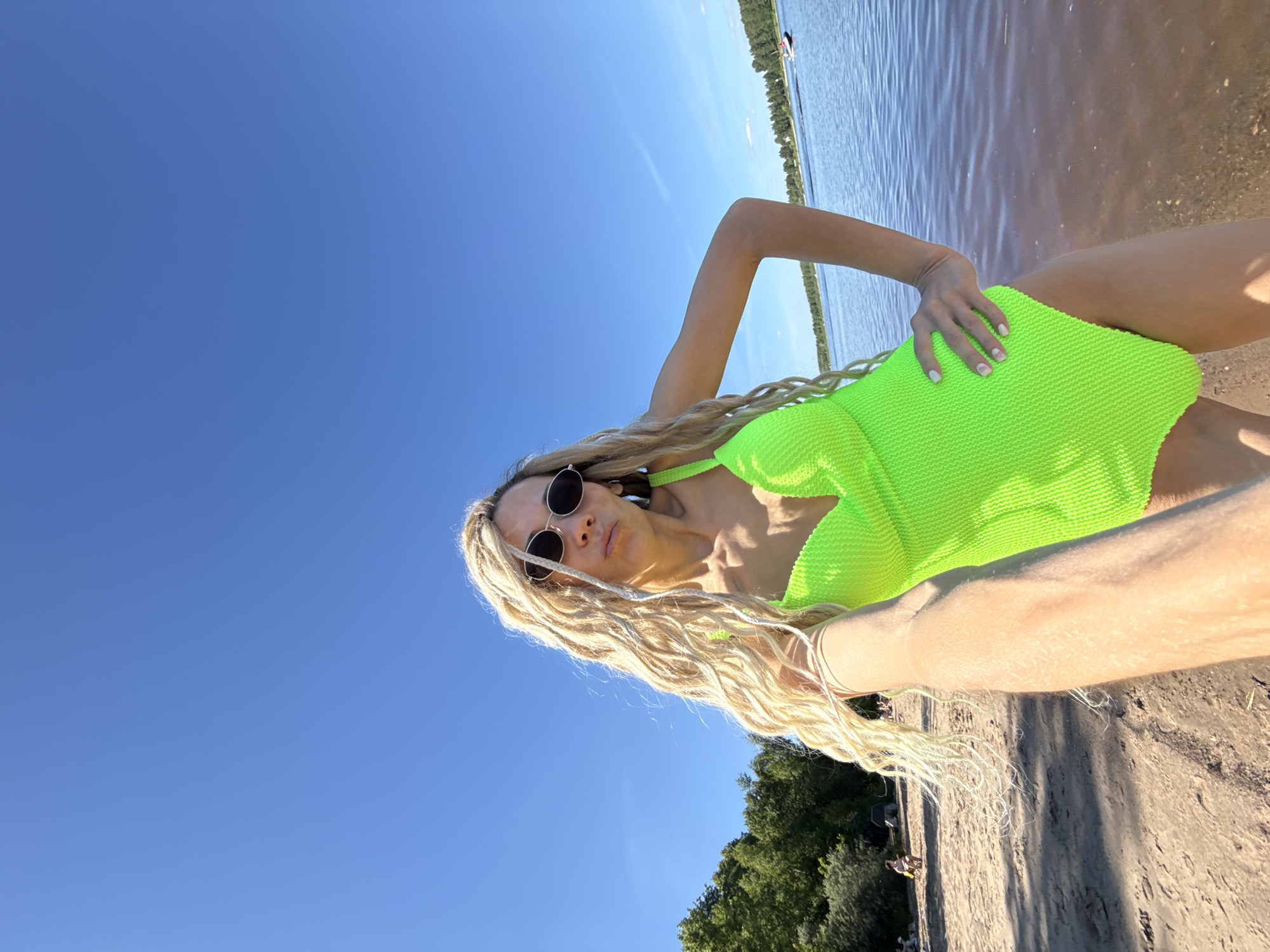 A female subject poses for a selfie on a sunny lakeside or riverside beach in Rusanovo, Russia. The woman, with long blonde dreadlocks and dark oval sunglasses, wears a textured neon green one-piece swimsuit. She faces the viewer directly, her right arm extended presumably holding the camera, and her left hand resting on her hip. Her lips are slightly pursed.
The scene is under a clear, bright blue sky devoid of clouds. A large body of water, appearing bluish-brown near the sandy shore, extends into the background, where a white jet ski with an operator creates a small wake. The far bank of the water is lined with dense green trees. On the sandy, light brown beach to the left, a few indistinct figures are visible relaxing. Lush green trees are also present on the left side of the near bank. The bright daylight casts gentle shadows on the sand.