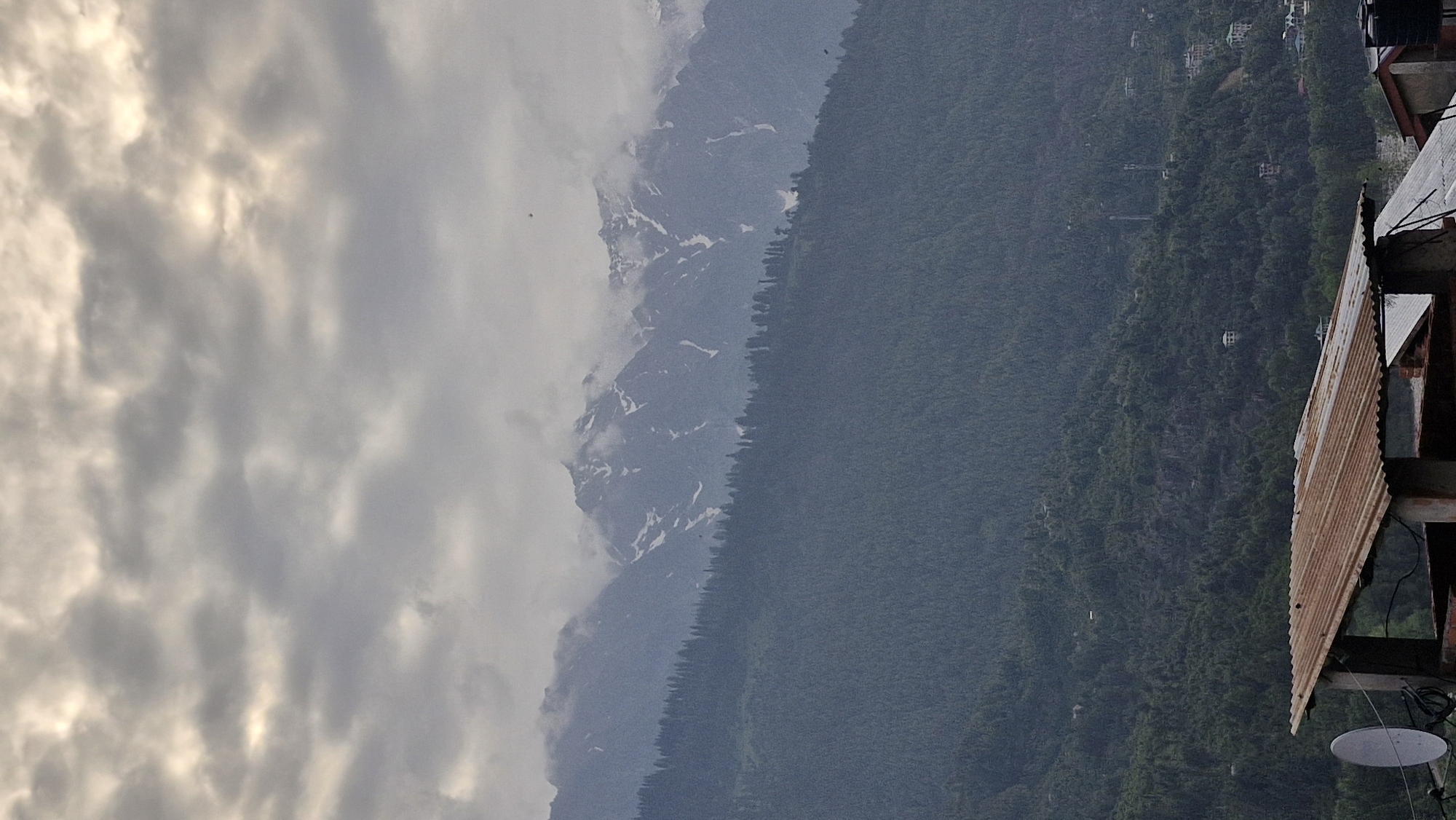 The image captures a panoramic view of the Vashisht region in India. The sky is overcast with a layer of gray clouds interspersed with patches of sunlight. In the background, majestic snow-capped mountains rise, their peaks partially obscured by mist. A dense forest covers the lower slopes of the mountains, transitioning into more sparsely vegetated hills closer to the foreground. In the lower section of the image, a rustic structure with a corrugated iron roof is visible, featuring a satellite dish and exposed brickwork. The overall scene depicts a tranquil, mountainous landscape with hints of human settlement.