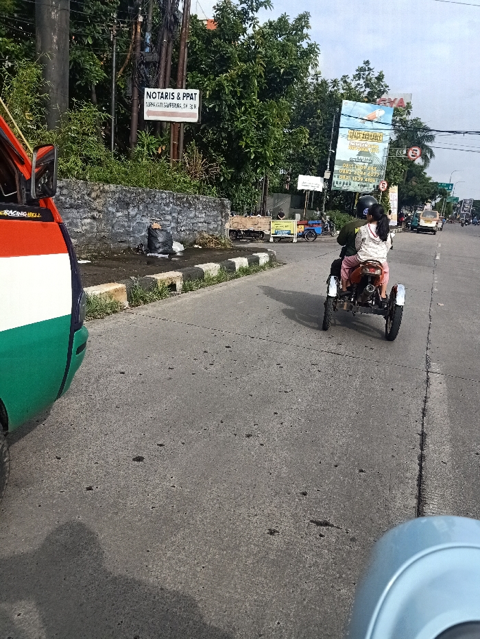 A motorized three-wheeled vehicle travels down a grey asphalt road. It carries two people, with the rear passenger wearing a patterned jacket and a helmet. The vehicle casts a shadow on the road. To the left, a colorful van is partially visible. In the background, lush green trees line the road, punctuated by signs, including one for "NOTARIS & PPAT." Other vehicles are further down the road. A blue object appears at the bottom right corner.