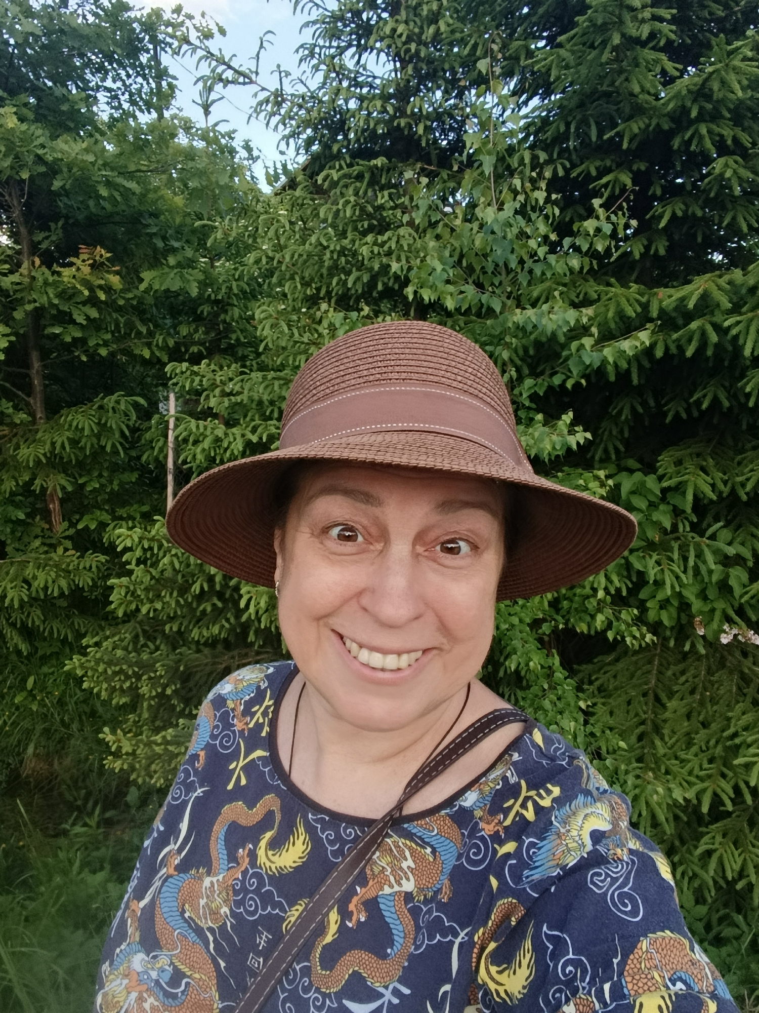A close-up outdoor portrait captures a smiling woman looking directly at the viewer. She has light skin, dark eyes, and a joyful expression, with a wide, toothy smile. She is wearing a brown woven straw hat with a darker brown ribbon band. Her top is dark blue or navy, adorned with an elaborate pattern of orange, blue, and yellow dragons, stylized clouds, and decorative graphic elements resembling Asian characters. A brown strap, likely from a bag, is visible over her right shoulder.
The background is filled with lush, dense green foliage, featuring a mix of deciduous trees with broad leaves and coniferous trees with needle-like branches. A small patch of light blue sky is visible through the canopy at the top. The lighting suggests it is daytime. The scene conveys a pleasant, natural outdoor setting, possibly in a garden or forest edge in Vereya, Russia. The woman appears happy and engaging.