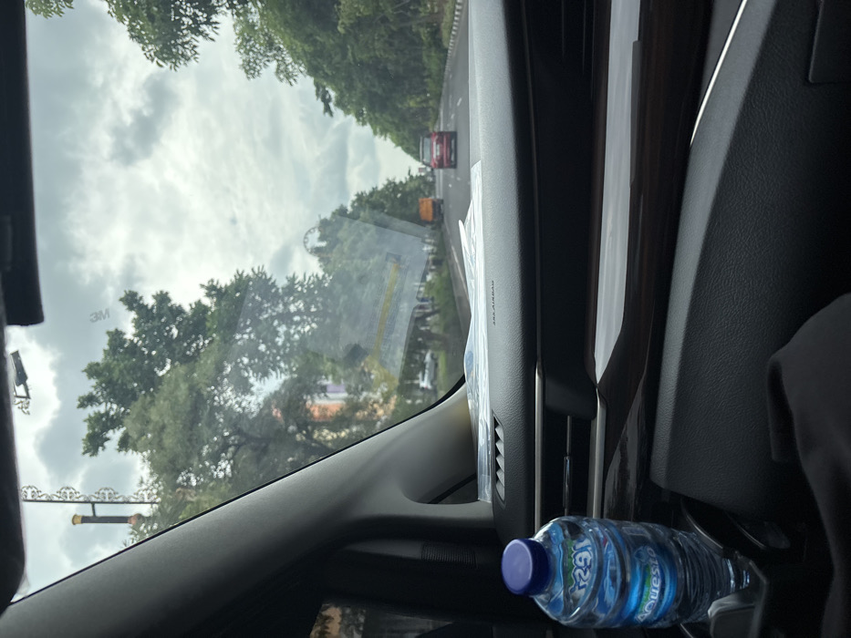 The image captures a view from inside a car, looking out the front windshield on an overcast day in Surabaya, Indonesia. The sky, filled with soft, diffused clouds, dominates the upper part of the scene. Lush green trees line both sides of the road ahead, creating a canopy effect.
On the road, a red car is visible in the distance, followed by what appears to be a yellow or orange vehicle, suggesting traffic. To the lower left, a decorative metal structure, possibly a street light or signpost, can be seen partially.
Inside the vehicle, the dark-toned dashboard and air vents are prominent in the lower and right sections of the frame. A label reading "AIRBAG" is visible on the passenger side of the dashboard. On the windshield, a small sticker with "3M" is discernable. A plastic water bottle, with "AQUA" (reversed) and "1624" printed on its label, rests on the dashboard near the center console. The car interior appears modern and well-maintained. No people are visible in the image. The scene suggests travel during daylight hours, under cloudy weather conditions.