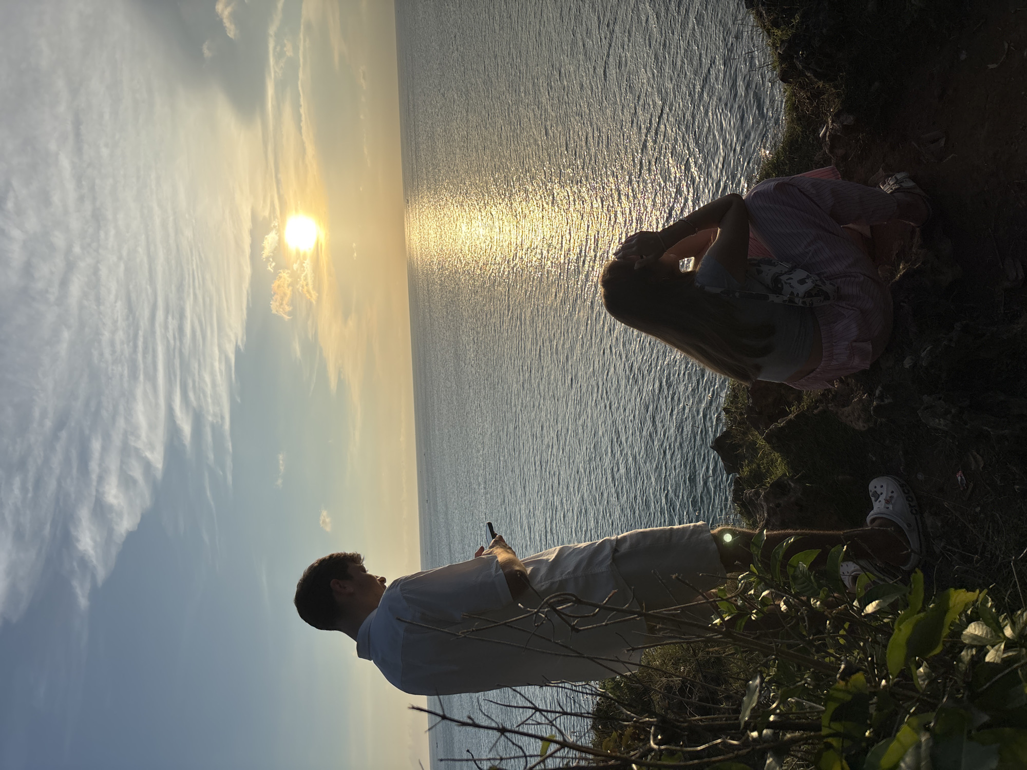 The image depicts two individuals at Uluwatu, Kuta Selatan, Bali, Indonesia, with the sunset over the ocean as a backdrop. One person is standing and appears to be looking at their phone. The other person is sitting, with their head in their hands. The environment consists of the ocean, sky, and cliffside vegetation.