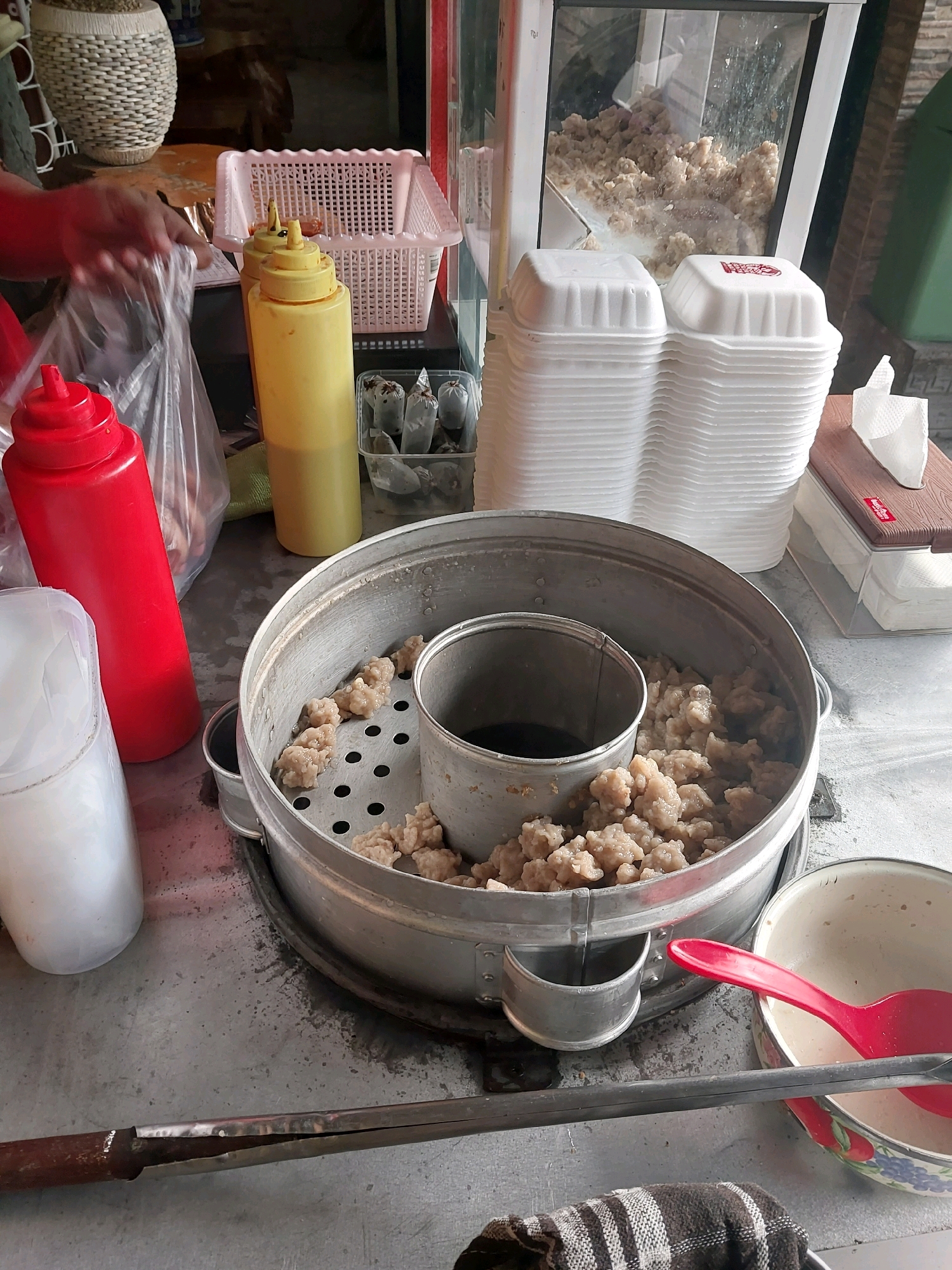 The image depicts a street food stall in Malang, Indonesia, likely selling meatballs (bakso) or a similar dish. In the foreground, a large aluminum steamer pot is filled with small, light-brown, round food items, presumably meatballs, arranged on a perforated tray. The steamer has a central cylinder containing a dark liquid, possibly broth or sauce. To the left of the steamer are two large squeeze bottles, one red and one yellow, commonly used for condiments like chili sauce and mustard. Next to the red bottle is a clear plastic bag containing more food items. To the right of the steamer is a white styrofoam bowl with a red plastic spoon inside. Further to the right are stacks of white styrofoam containers for takeout. In the background, a person's hands are visible, holding a clear plastic bag, suggesting preparation or packaging of food. A white woven basket and a white plastic basket containing some items are also present in the background. The stall appears to be set up outdoors or in an open-air market, with a tiled surface beneath the displayed items. The overall scene is one of food preparation and service at a local eatery.