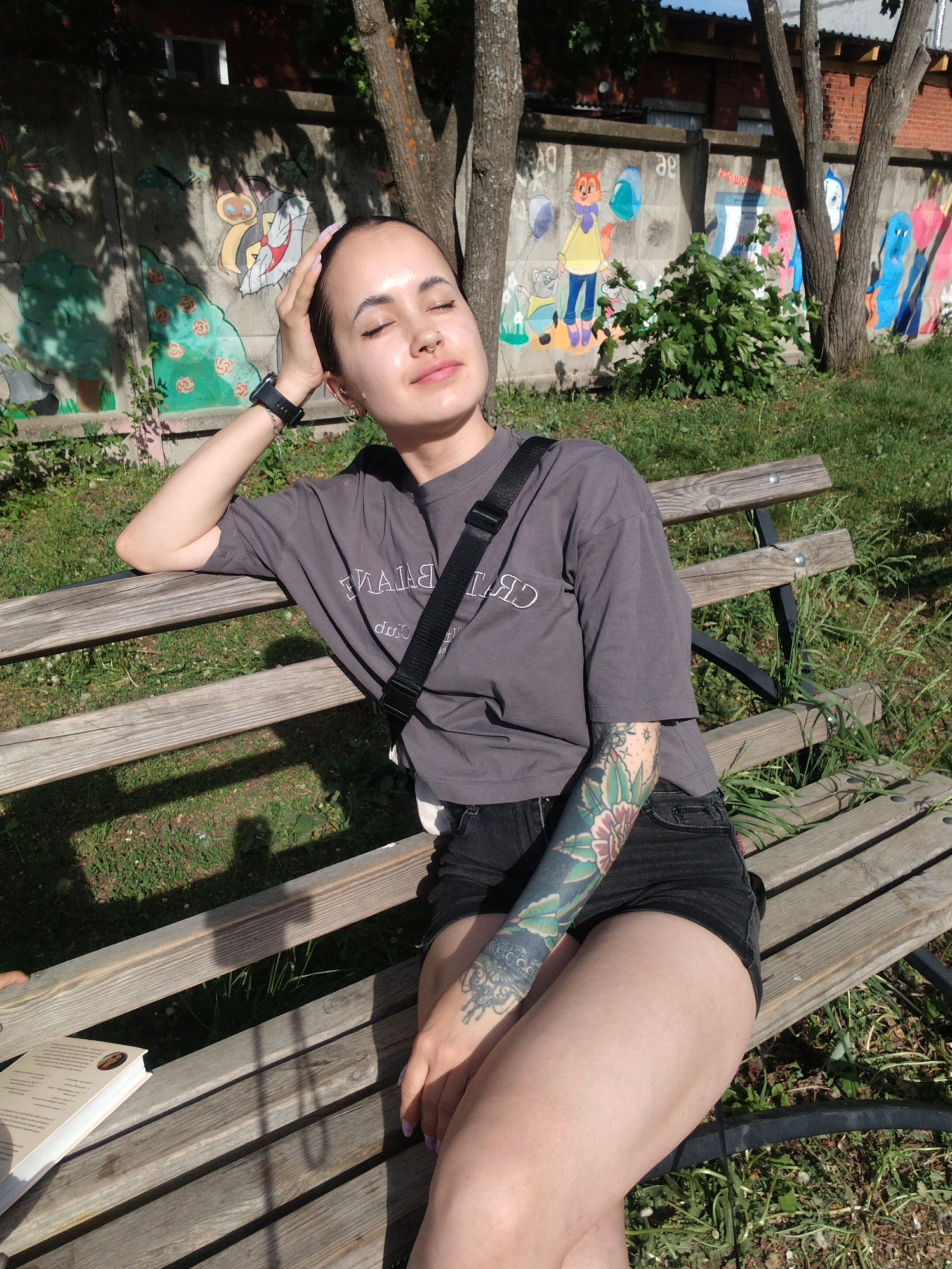 A woman is sitting on a park bench in Noviy, Russia, soaking up the sun. She has her eyes closed and her head is tilted back, as if to better feel the warmth on her face. She's wearing a gray t-shirt and black shorts. A black strap runs across her torso, suggesting she might be carrying a bag. The woman has a tattoo that covers her left arm. She is resting her left hand on her head. A wristwatch and a bracelet are on her left wrist. A book sits on the bench near her left hip. Behind the bench is a concrete wall covered in colorful murals. Several trees are visible in the background.