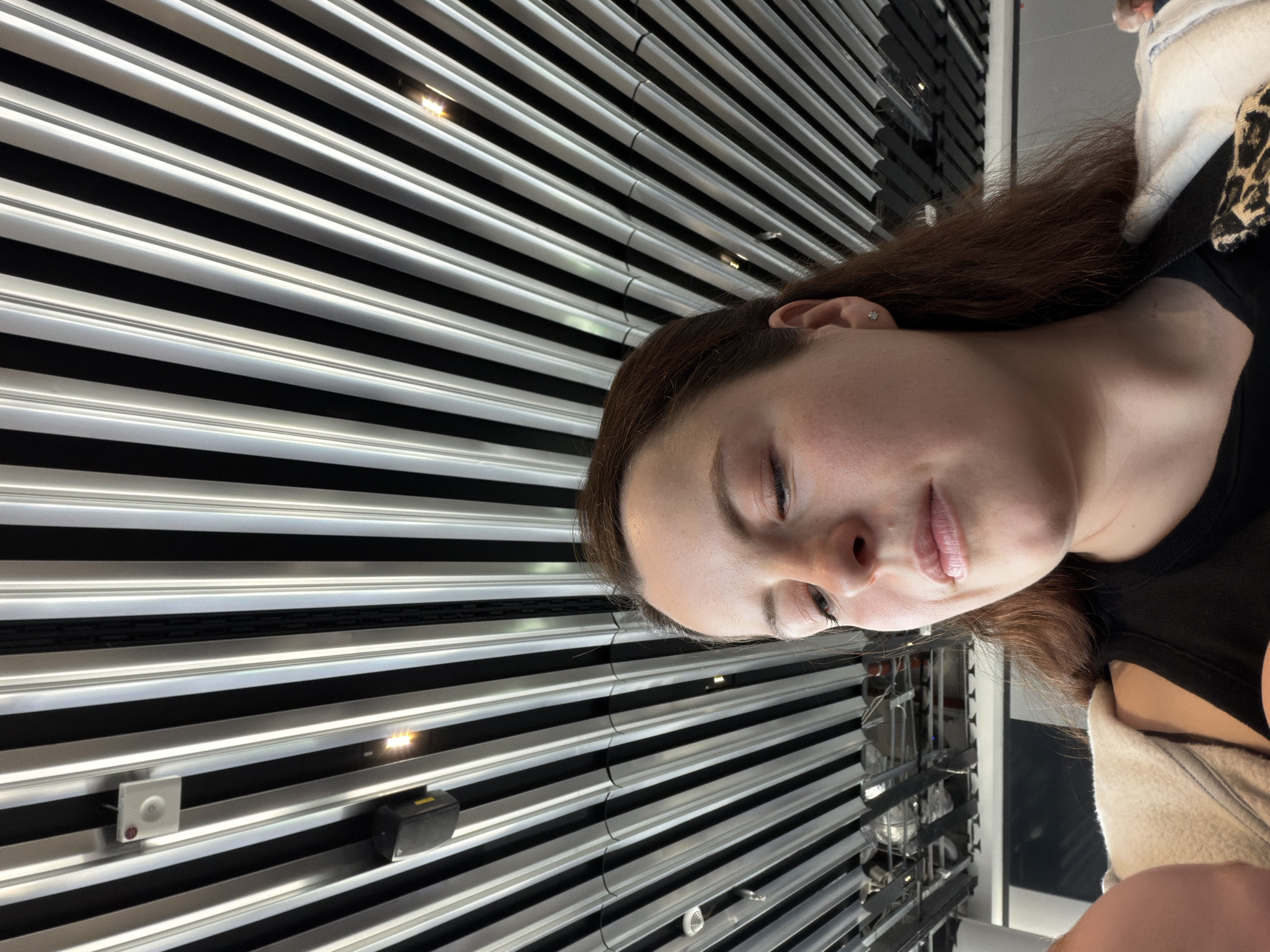 The image features a person with closed eyes, light skin, and brown hair. They are wearing a black top with a beige jacket visible at the edges. The background is dominated by a linear pattern of horizontal silver and black stripes, possibly a ceiling or wall structure. In the lower right corner, there are glimpses of a complex metallic structure, potentially part of the building's infrastructure or equipment.
Given the architectural style and the presence of a sophisticated building infrastructure, it's plausible that this image was taken inside a modern building in Frankfurt, Germany. Specific confirmation would require additional visual cues or contextual information.