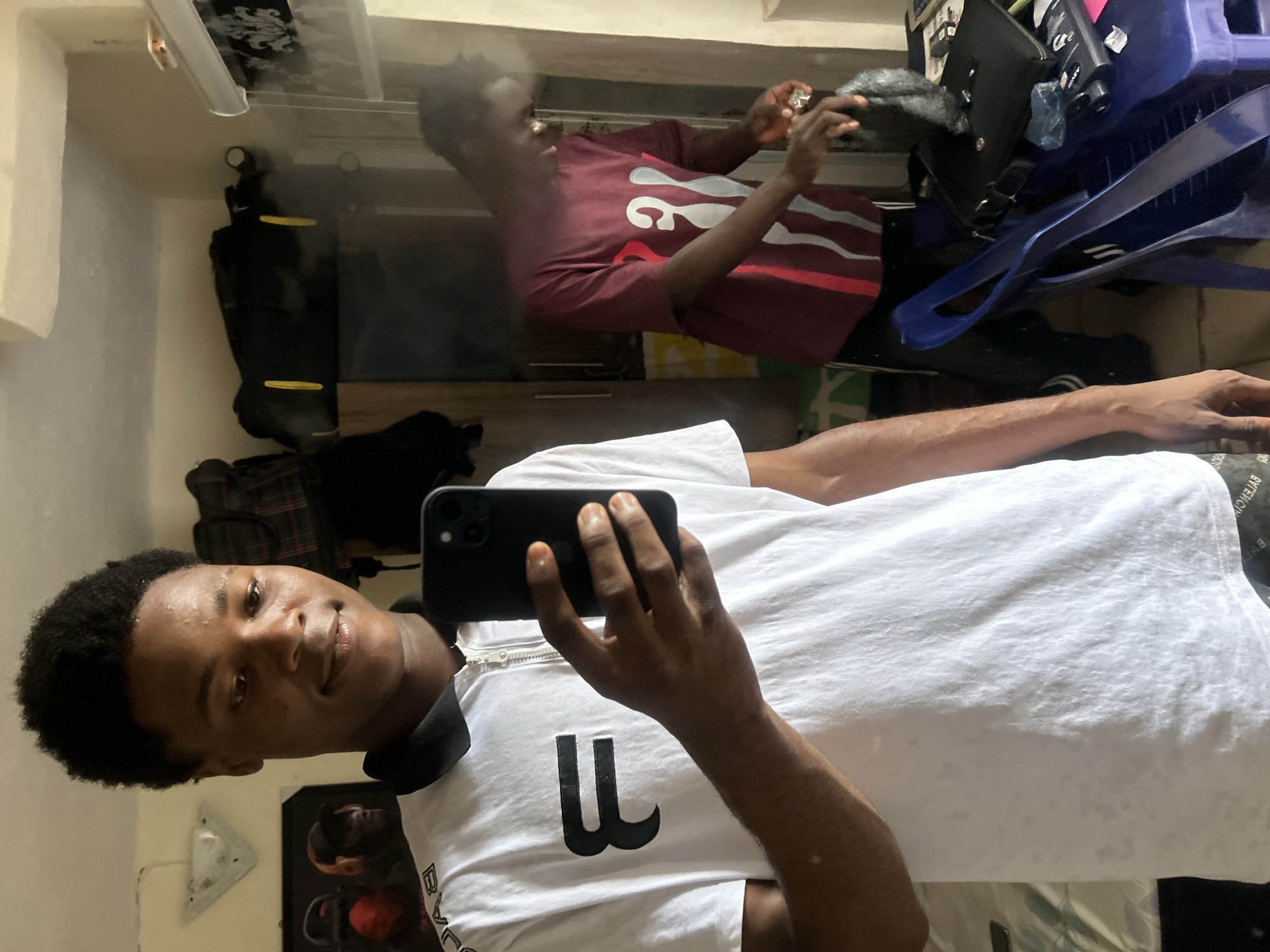 The image depicts an indoor scene in what appears to be a residential room in Ihiagwa, Nigeria. A young male, approximately in his late teens or early twenties, with dark skin and short dark kinky hair, is positioned in the foreground, taking a mirror selfie. He is wearing a white short-sleeved polo shirt with a black collar, a zippered placket, and a large black stylized "F" or "B" symbol on the chest. The word "BALENCIAGA" is visible in reverse print along the lower edge of what appears to be his shorts or trousers. He holds a black smartphone with multiple rear cameras in his right hand, pointed towards himself.
Reflected in the mirror, another young male of similar age and complexion is visible in the background, standing to the right. He is wearing a maroon short-sleeved t-shirt with vertical white and light grey abstract patterns on the front, and dark trousers. He appears to be engaged in handling a black object, possibly a bag, with both hands on a blue plastic stool or small table.
The room features light-colored walls, possibly cream or beige. A triangular-shaped light fixture is mounted on the upper left wall. Behind the foreground individual, a framed picture or poster with two figures, possibly wearing hats and headphones, is partially visible. To the right of this, a dark wooden cabinet or wardrobe is present, topped by a black duffel bag with yellow accents. Another dark garment bag hangs from the side of the cabinet. On the blue plastic stool/table in the background, a black structured bag (possibly a briefcase or messenger bag), a black rectangular object (possibly an electronic device), and some scattered papers are laid out. A patterned blind or curtain with a black and white/grey floral design is visible on the far right. The floor appears to be a light, possibly tiled, surface.