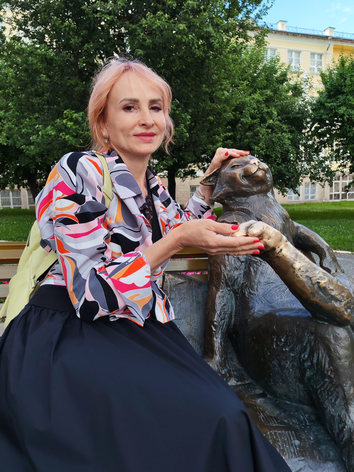 A middle-aged woman with pink hair, wearing a colorful geometric-print jacket over a black top and a dark skirt, is seated on a bench next to a bronze statue of a cat in a park in Yoshkar-Ola, Russia. The woman is gently stroking the cat's head with her right hand and holding its paw with her left hand. She has red nail polish on. The statue of the cat is depicted in a relaxed, reclining pose. The background shows green trees and a building with yellow walls and white windows, suggesting an urban park setting. The sky is partly cloudy.