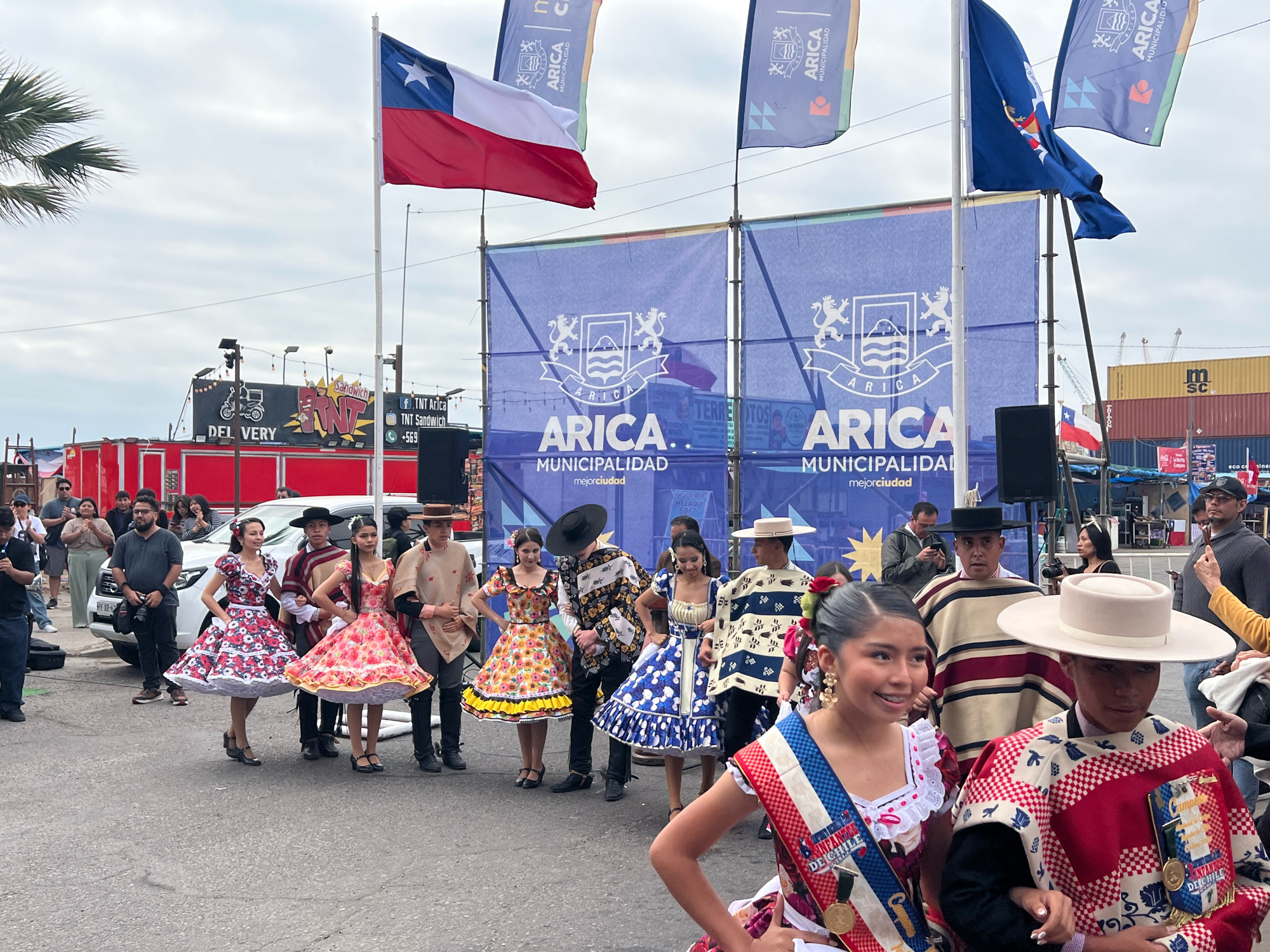 The image depicts an outdoor street scene in Arica, Chile, likely during a cultural event. In the foreground, several young people are dressed in traditional Chilean attire. Two young women wear brightly colored, floral-patterned dresses with full skirts and cinched waists. They are accompanied by young men in ponchos and wide-brimmed hats. One young woman in the immediate foreground wears a sash with "Infancia de Chile" printed on it. Behind them, a white SUV is parked on the street.
In the mid-ground, a large blue banner with the text "ARICA MUNICIPALIDAD" is prominently displayed, featuring the city's coat of arms. Flags are also visible, including the Chilean flag flying from a pole, and other flags with the "ARICA MUNICIPALIDAD" logo. The background shows a cityscape with buildings, shipping containers, and cranes, suggesting a port area. The sky is overcast. People are gathered, some watching the procession or event, while others appear to be participants. The overall atmosphere suggests a festive or ceremonial gathering in the city.