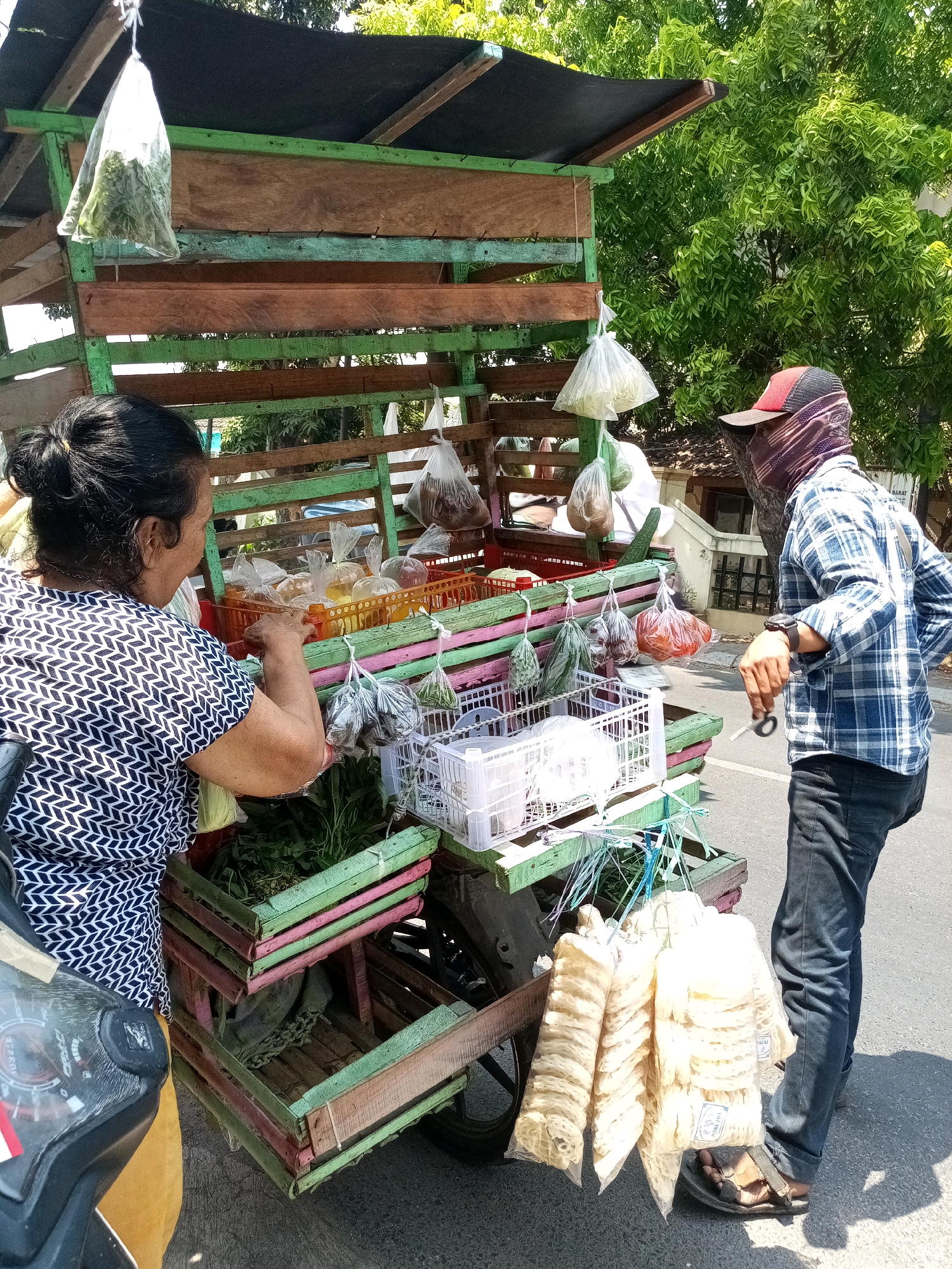 In Bekasi, Indonesia, a street vendor operates a mobile cart laden with produce and snacks. The cart, constructed from weathered wood and painted in vibrant green and pink, is laden with various goods. Bags of leafy greens hang from the top, while plastic-wrapped vegetables and fruits are arranged in orange baskets on the middle shelves. Below, a white plastic crate holds more greens. The lower section of the cart reveals wooden crates, one with a sack inside and another empty. Hanging from the side of the cart are multiple packages of what appear to be dried crackers or chips.
Two individuals are interacting with the cart. A woman, with her hair tied back, stands on the left, wearing a black and white patterned shirt. She is reaching into one of the baskets, selecting items. On the right, a man wearing a blue plaid shirt, dark pants, a red and black cap, and a patterned face covering is observing. He has his left hand in his pocket and appears to be holding a small object, possibly scissors, in his right hand. A motorcycle is partially visible in the foreground on the left. The scene is bathed in bright sunlight, casting distinct shadows, suggesting it is a clear day. The background shows lush green trees and parts of buildings.