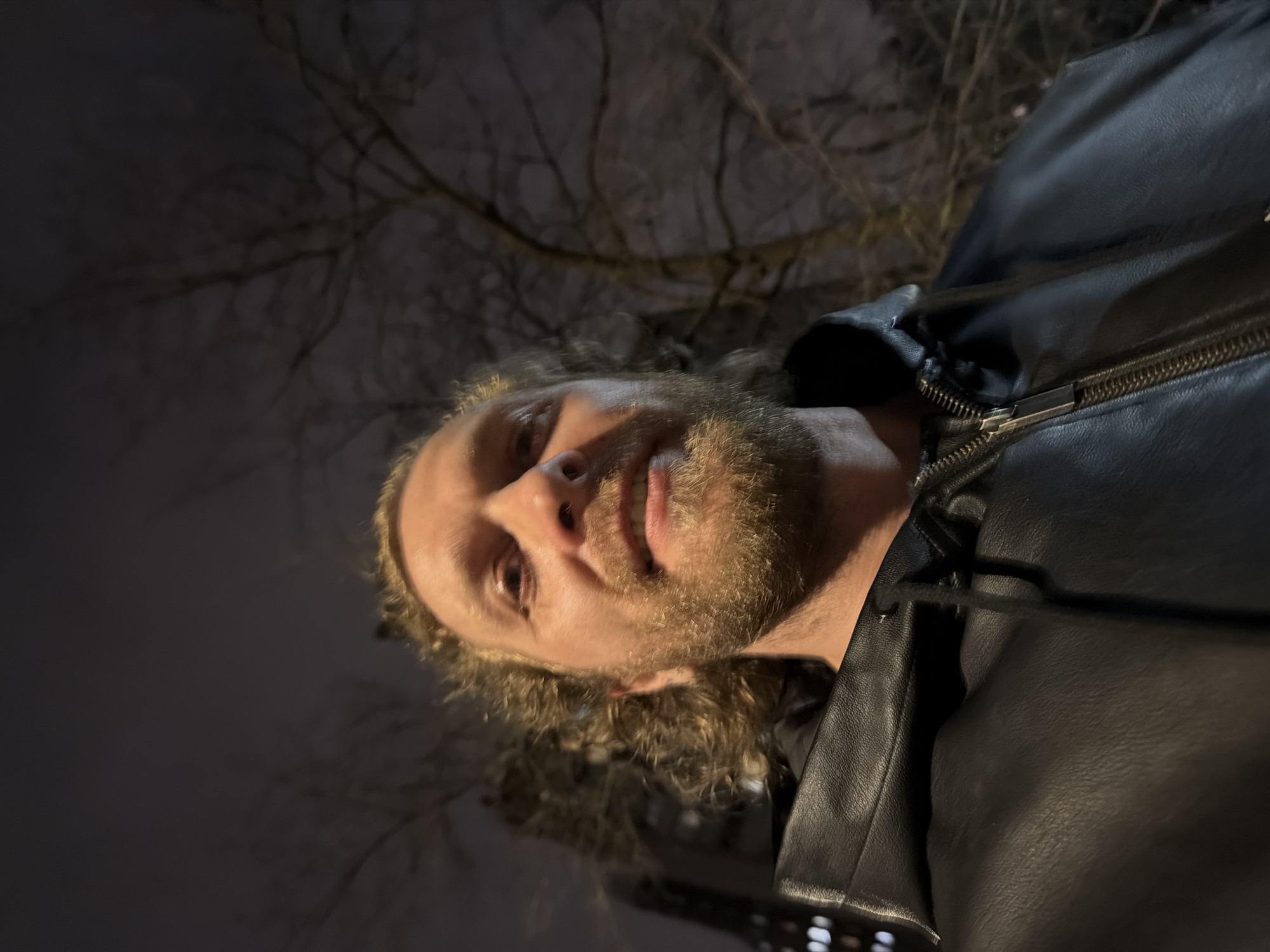 The image depicts an adult male captured in a low-angle selfie at night in Saint Petersburg, Russia. The man has light-colored, curly hair and a full beard, and displays a slight, closed-mouth smile. He is wearing a black hooded leather jacket with a visible zipper and drawstring. The background features bare tree branches against a dark sky in the upper right quadrant, suggesting a deciduous tree. In the upper left background, a blurry, multi-story building with illuminated windows arranged in a grid pattern is partially visible. The scene is illuminated by artificial light, casting a glow on the man's face from below and creating shadows under his chin. No specific street or prominent landmark within Saint Petersburg is discernible.