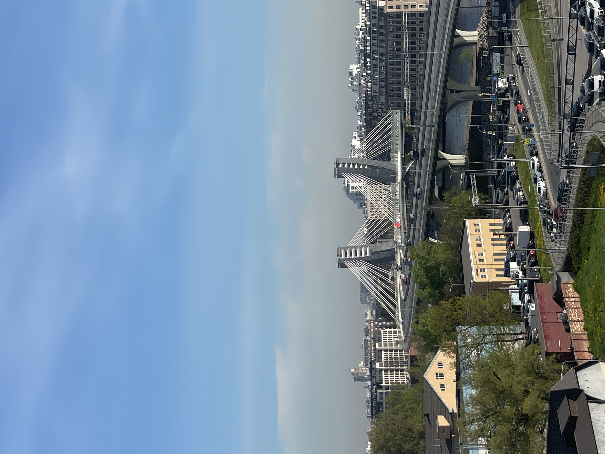 The image depicts a city scene featuring modern architecture, infrastructure, and a sky with varying degrees of cloud cover. Prominent features include suspension bridges with white cables, elevated highways, and dense urban development with high-rise buildings. There is heavy traffic congestion on the roads. A mix of commercial and residential buildings are visible along with green spaces containing trees and grass.