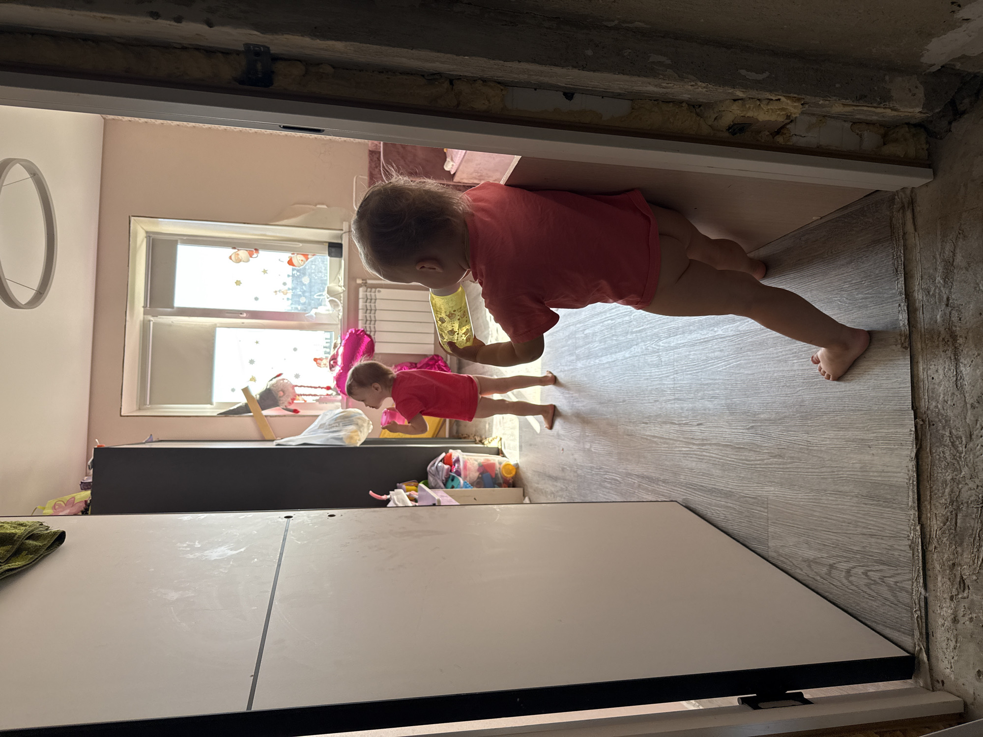 A vertically oriented photograph taken indoors shows two young children in a room that appears to be undergoing renovation. In the foreground, a white cabinet dominates the bottom half of the frame. Behind it, a dark cabinet is visible, stocked with various toys in a clear plastic bin.
To the right of the dark cabinet, a young child, dressed in a pink t-shirt and shorts, is standing and holding a yellow cup to their mouth. Further to the left and slightly behind this child, another child, also in pink, is lying on the floor.
The room has light pink walls and a window with a view of an urban setting, suggesting it's daytime. The floor is covered in wood-look laminate. The ceiling area above the doorway shows exposed insulation and rough concrete, indicating unfinished construction. The overall scene suggests a domestic setting in Chelyabinsk, Russia, where everyday life continues amidst renovation work.