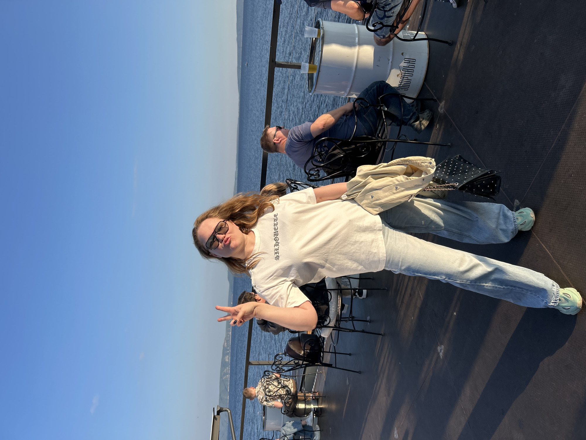 A young woman with blonde hair and sunglasses poses for a photo on a boat, holding up two fingers in a peace sign. She is wearing a white t-shirt, light-wash jeans, and light blue sneakers. Next to her is a black studded purse and a light-colored jacket. In the background, two men are seated on a boat deck, looking out at the water. One man, wearing a blue t-shirt and jeans, is reclining in his chair. Another man, in a patterned shirt, is seated further back. A white barrel with two glasses of amber liquid on it serves as a table. The scene is set on a sunny day, with a clear blue sky and calm water. The boat appears to be on a body of water with hills or land visible in the distance. Text on the white barrel is partially visible and appears to be in Cyrillic, possibly related to a brand or location.