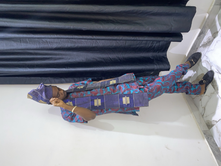 A man in vibrant blue and red patterned Yoruba attire is seen reclining. He wears a matching cap and suede loafers. A sash with blue and metallic patterns adorns his upper body. A dark, draped curtain forms a backdrop. The setting is indoors, suggested by a white wall and marble-tiled corner.