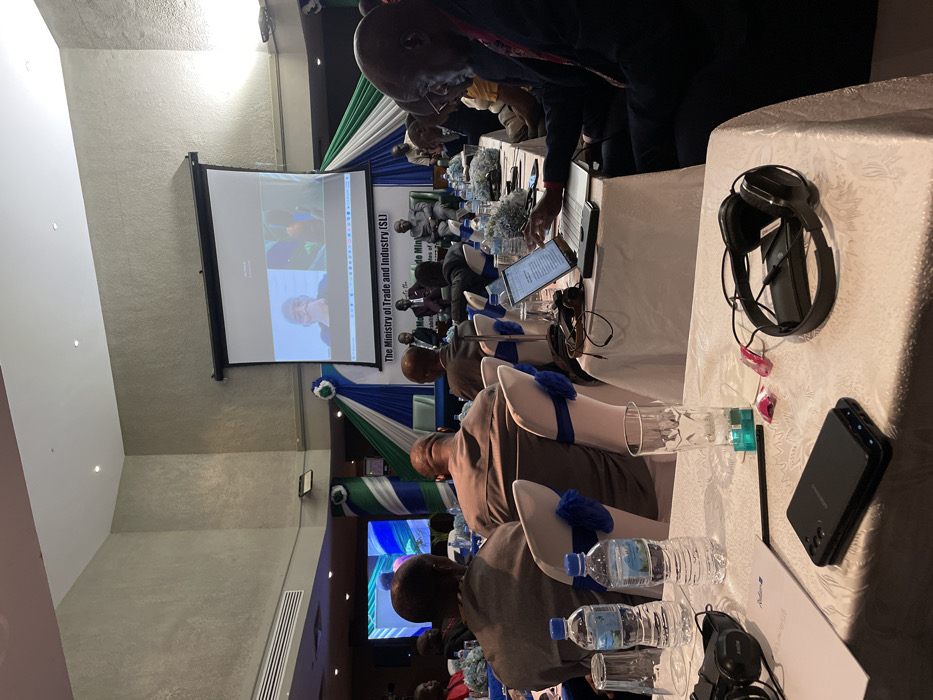 This image captures an indoor meeting or conference taking place in Freetown, Sierra Leone, likely during the daytime under artificial lighting. The scene shows multiple individuals, predominantly men, seated at long tables covered with white tablecloths. The chairs are adorned with white covers and distinct blue bows, matching blue decorative elements on the tables.
In the background, a large projector screen displays a video call, showing two individuals, indicating remote participation or a hybrid event setup. Below the screen, a banner is visible with the prominent text: "The Ministry of Trade and Industry (SL)". Partially visible text beneath this includes "Directorate of" and "Ministers of", strongly suggesting a government or ministerial meeting related to trade and industry in Sierra Leone. The draped fabric in the background features blue, white, and green colors, consistent with Sierra Leone's national flag.
Attendees are seated facing the front of the room, with some using laptops or tablets placed on the tables. Each place setting includes bottled water, clear glasses, and in some instances, headphones, implying simultaneous translation or audio equipment. In the foreground, on a table to the right, a pair of black headphones and a black smartphone are visible, along with another water bottle and a clear glass. A document with "Radisson Blu" is also faintly seen on one of the tables, possibly indicating the venue or a sponsor. The atmosphere appears professional and focused, with participants engaged in the proceedings of the event.
