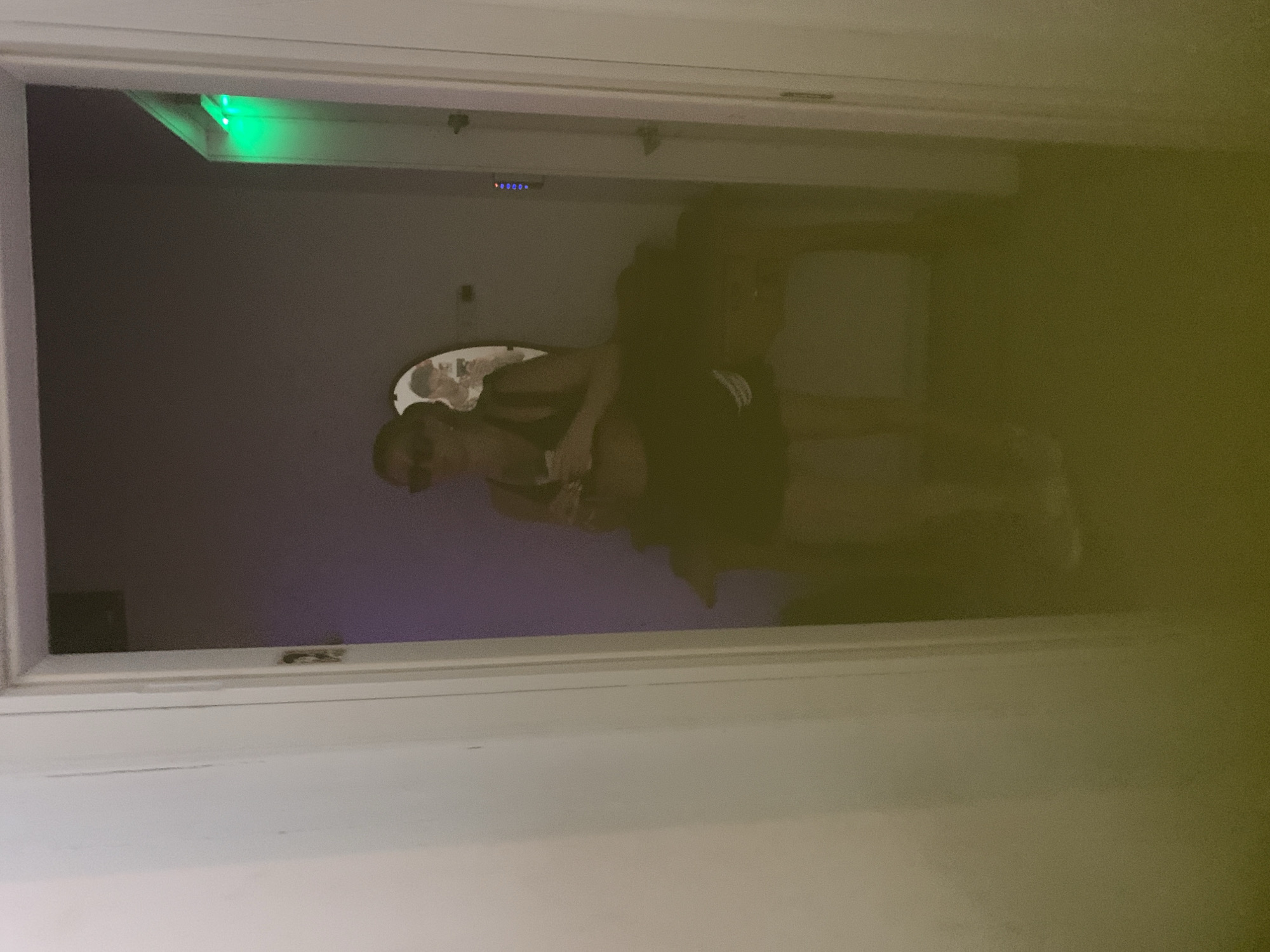 The image shows an interior view through a white door frame. Inside the room, a person is visible, wearing sunglasses and dark clothing. The room appears to be dimly lit, with a purplish hue in the background. There is a small mirror on the wall. A strip of green LED lights is affixed to the top of the door frame. There is no information available in the image to ascertain the location in the city of Tel-Aviv, Occupied Palestine.