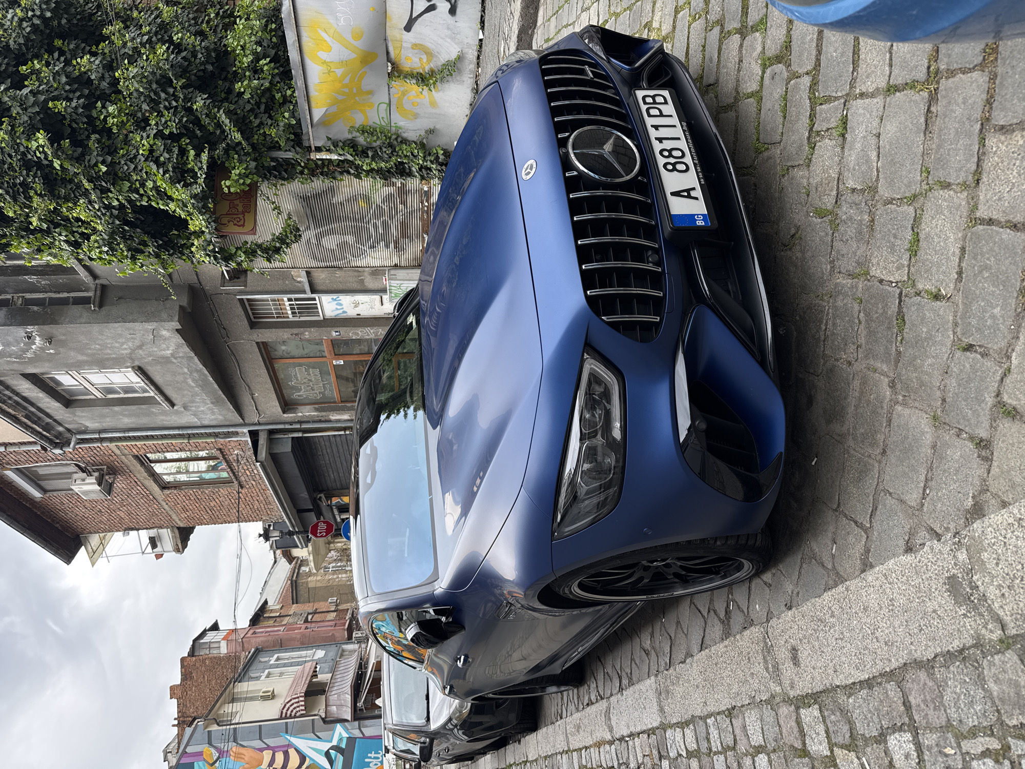 A blue Mercedes-Benz, likely an AMG model with a satin or matte finish, is parked on a cobblestone street in Plovdiv, Bulgaria. The front of the car is angled towards the viewer, showcasing its prominent grille with vertical slats and the Mercedes-Benz star emblem. Sleek headlights and black multi-spoke wheels are visible. The European license plate, positioned on the front bumper, reads "A 8811 PB", includes the country code "BG", and has "www.autohaus-bg.com" printed on its frame.
The street is paved with old, grey cobblestones, with small patches of green grass growing in the cracks, indicative of an older urban setting. A concrete curb separates the roadway from the sidewalk. In the background, a smaller, dark car is partially visible.
The surrounding architecture features a mix of older buildings. On the left, a wall is extensively covered in lush green ivy, beneath which a small, partially obscured red sign can be seen. Above the ivy, yellow spray paint graffiti mars a corrugated metal surface. Other buildings feature exposed brickwork, painted facades, windows, and air conditioning units. A red octagonal "STOP" sign is visible on a pole further down the street. Graffiti in blue and white is present on a lower wall in the background, adjacent to a more colorful mural. The sky is overcast, casting a soft, diffused light over the scene, suggesting a cloudy day. No people or active events are captured in the frame.