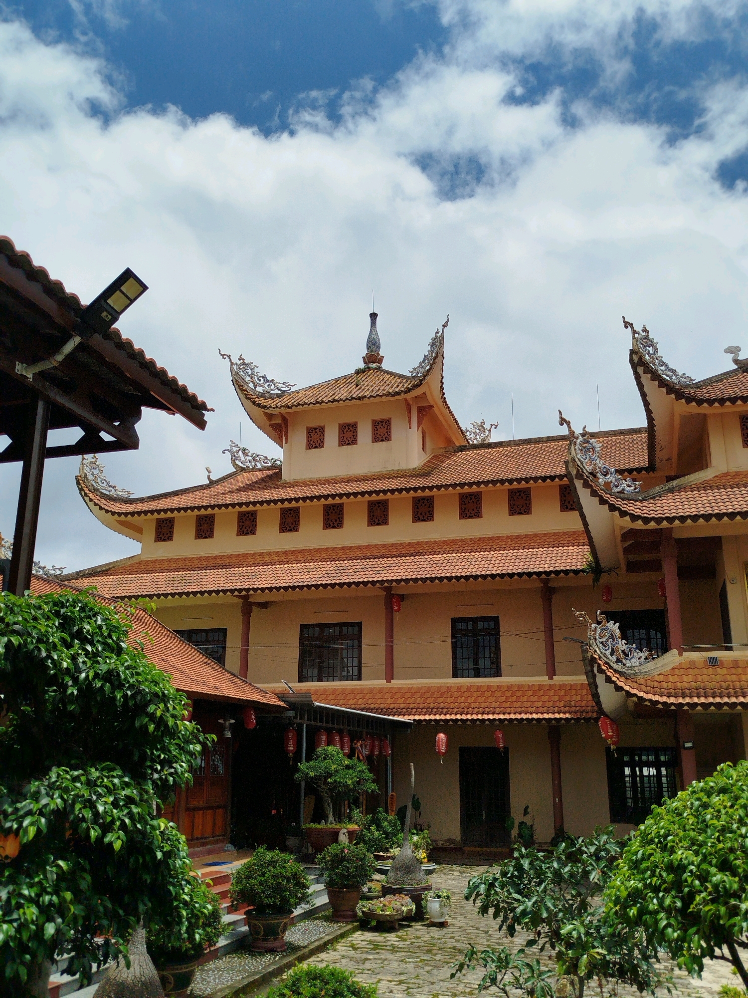 The image shows a courtyard of a Buddhist temple, likely in Lâm Viên - Đà Lạt, Vietnam, due to the distinct architectural style. The main building, painted a light yellow, features a multi-tiered, curved tiled roof with ornate dragon-like decorations on the eaves. Several windows with intricate lattice patterns are visible on the upper floors. The courtyard is paved with stones and decorated with several potted bonsai trees and other lush greenery. Red lanterns hang from the eaves of the building and a covered walkway. The sky above is blue with scattered white clouds. A modern solar-powered light fixture is attached to a covered structure on the left side of the frame. No people are visible in the image.
