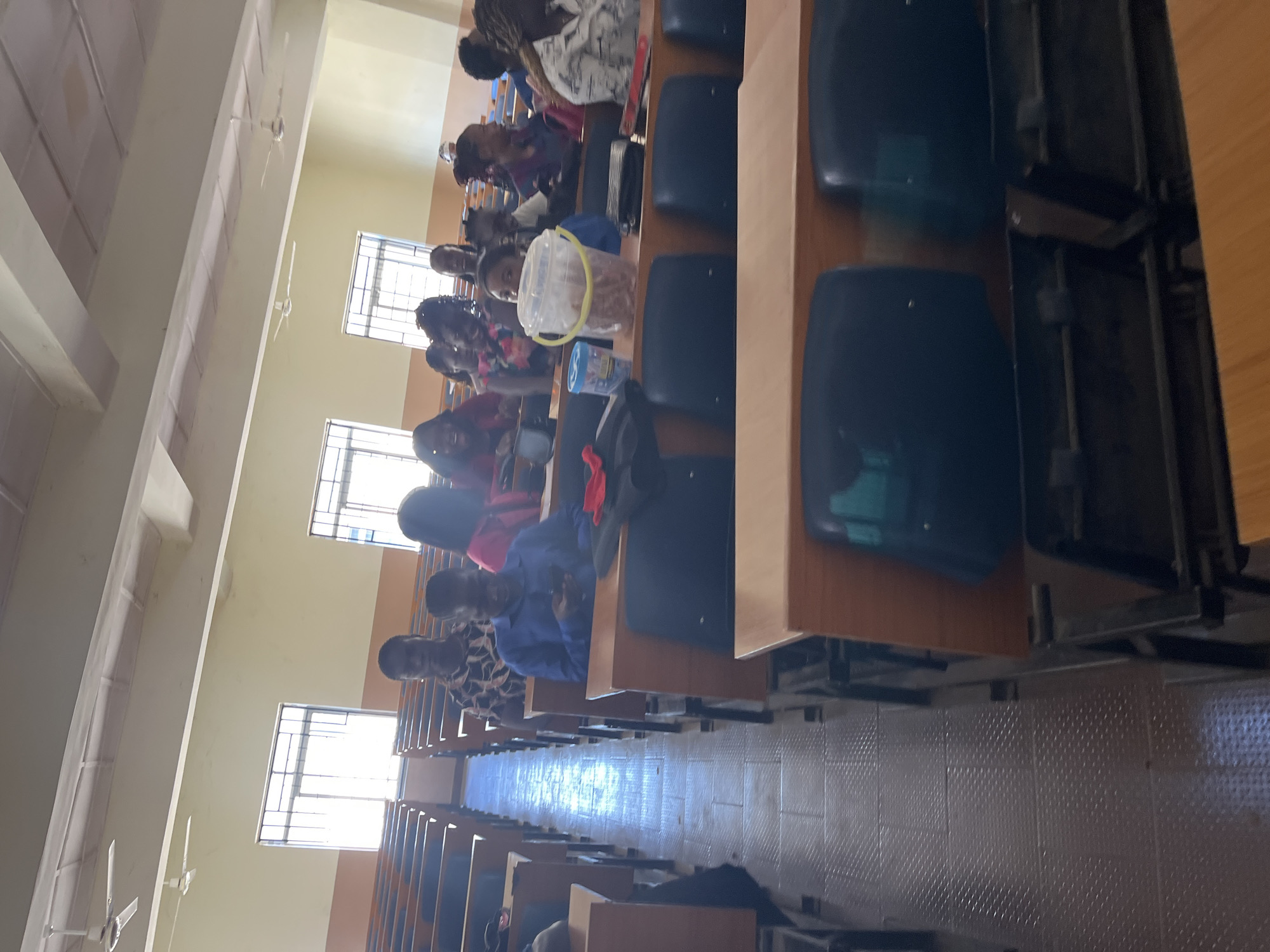 The image depicts an indoor scene, likely a lecture hall, in Jos, Nigeria. Several individuals, appearing to be students, are seated at desks arranged in rows. The room is illuminated by overhead lighting, and ceiling fans are visible, suggesting a warm climate.
In the foreground, rows of wooden desks with dark seats are prominent. The floor appears to be tiled with a textured pattern.
Several individuals are visible in the mid-ground and background, seated and facing forward. Some are looking at their phones, while others appear to be engaged with their surroundings. One person in the mid-ground is wearing a blue shirt and looking down at their mobile phone. Another individual to their right is wearing a patterned shirt. Further back, several women are seated, some with dark hair.
On one of the desks in the mid-ground, a transparent bucket filled with a light-colored substance and a blue container are present. There are also dark bags and some red fabric on some of the desks.
The overall environment suggests an academic setting, possibly a university or college in Jos, Nigeria, during a lecture or study session. The presence of ceiling fans and the natural light from the windows indicates daytime.
