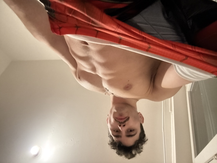 This indoor image captures a young adult male from a low-angle perspective, with the subject appearing upside down. The man, positioned centrally, has dark, curly hair and a short mustache, and he looks directly at the camera with a neutral expression. A thin necklace with a small pendant is visible around his neck.
His upper torso, including his chest and defined abdominal muscles, is prominently featured. He is partially dressed, with a red and black patterned garment, possibly a shirt or top with a design resembling a spiderweb, pulled upwards, revealing his bare midsection and a pair of white briefs underneath. Further up, a grey garment, likely another layer of underwear or shorts, is also partially visible beneath the red and black clothing.
The setting is a simple indoor room with light-colored walls or ceiling. A bright, artificial light source, possibly a recessed light fixture, is visible on the lower left side of the frame. On the right, a vertical white structure, possibly part of a door frame or room corner, can be seen. The lighting suggests an indoor environment, making the time of day indeterminate. There is no visible text in the image.