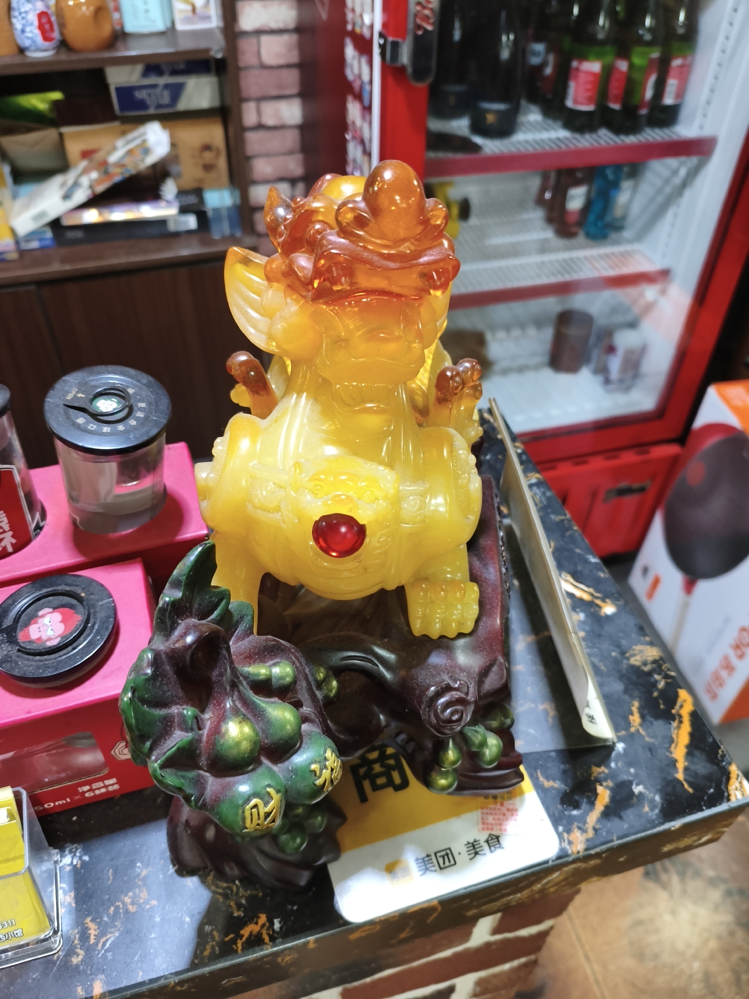 A close-up, eye-level shot captures a decorative statue of a mythical creature, likely a Pixiu or Qilin, made of translucent yellow material, possibly resin or amber, with orange translucent accents on its head and paws. A red gem is set into the creature's chest. The statue is mounted on a dark, carved base resembling a gnarled tree branch. In front of the statue, to the left, is a dark green carving of gourds with gold Chinese characters inscribed on them.
The statue and carvings rest on a black and gold speckled surface, possibly a tabletop or counter. To the left of the statue are several small, cylindrical containers with black lids. One is clear and contains a grey substance, and another is red with a black lid and Chinese writing. Behind these containers, a portion of a red refrigerator is visible, with shelves containing bottles of drinks. In the background, shelves hold various items, including boxes and decorative vases. The overall scene appears to be an indoor setting, possibly a shop or a home. The image does not provide any specific location within Jixi, China, other than suggesting an indoor commercial or residential environment. There are no people visible in the image, and no discernible actions or interactions are taking place. The lighting is moderate, illuminating the statue and its surroundings.
