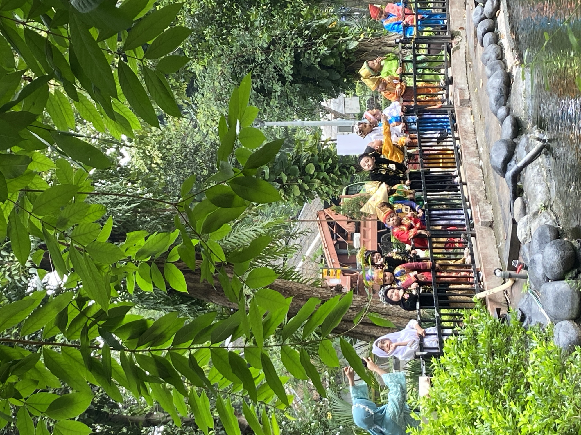 The original image is rotated 90 degrees counter-clockwise. When viewed upright, it depicts an outdoor cultural event in Surabaya, Indonesia, characterized by lush greenery and a water feature.
The central subjects are a group of young children lined up behind a black metal fence, which borders a shallow water body with a rocky edge and a small pipe extending into it. These children are dressed in vibrant, traditional Indonesian attire, featuring rich colors such as gold, red, blue, and green, often complemented by ornate hats or headbands. Their expressions and postures vary; some are actively observing something to their right, while others appear more relaxed, with one child in a black and gold outfit seemingly asleep or resting on the fence. The number "51" is visible on one child's garment.
In the foreground, large, verdant leaves from an overhead tree branch frame the scene. The background is filled with dense green foliage, through which a faint structure, possibly part of a building or an old vehicle, and a white utility pole can be discerned. Two adults are also visible: one in the lower foreground wearing a light blue traditional dress and hijab, extending an arm, and another in a white hijab standing further along the fence.
The scene unfolds during bright daytime under clear weather conditions, suggesting a school outing, cultural performance, or local festival. The overall atmosphere conveys a sense of vibrant cultural engagement within a serene natural setting.