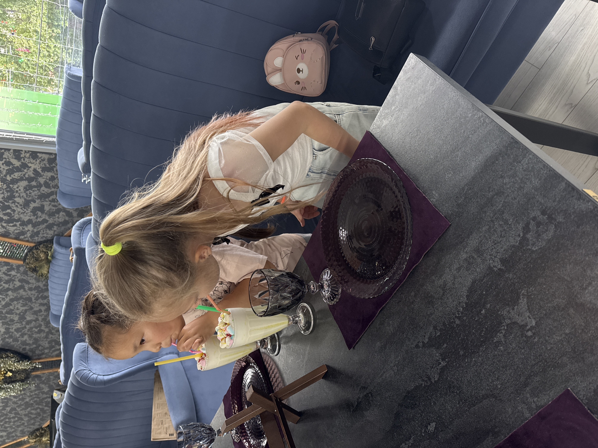 Two young girls are sitting at a table in an indoor establishment, likely a cafe or restaurant, in Anapa, Russia. The girl on the left, with dark hair in a bun, is drinking a milkshake with a yellow straw. The girl on the right, with blonde hair in a ponytail, is leaning over and also appears to be drinking a milkshake, possibly with a green straw. Both girls are wearing light-colored tops. On the table are two glasses filled with milkshakes topped with marshmallows, a dark glass, and two dark plates with purple placemats. A small pink backpack with a bunny design is visible on the blue upholstered bench seating behind the girl on the right. The background shows more blue seating and decorative elements. The floor appears to be light-colored wood laminate.
